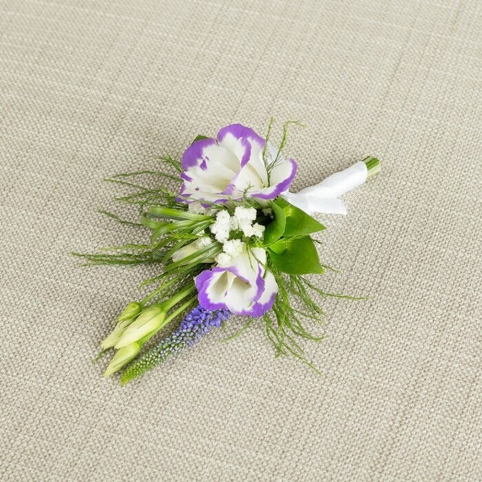 An elegant boutonniere is set against a softly textured light beige cloth, the fabric's fine weave and warm tone giving the impression of natural linen laid out on a dressing table before a special event. In the centre of this compact arrangement, two lisianthus flowers open in creamy white layers, each petal edged in a vivid, saturated purple that runs like ink along the ruffled contours, creating a striking yet refined two-tone effect. Their smooth, almost porcelain-like petals catch the gentle light in subtle highlights and shadows, adding depth and a sense of three-dimensionality. Around them, clusters of tiny white filler blooms-similar to gypsophila-provide a cloud-like backdrop, softening the contrasts and lending the piece a romantic, bridal feel that would suit a wedding at a local New Barnet church or registry office. Intertwined with these is a slim vertical stem of muscari, its deep purple, tightly packed, bell-shaped florets forming a textured column that contrasts beautifully with the lisianthus' open faces. Two pale green, unopened buds tuck into the side of the design, suggesting fresh growth and the promise of more flowers to come, reinforcing the sense of new beginnings. Fine, feather-textured greenery, likely asparagus fern or similar, fans out delicately around the blooms, its filigree fronds softening the outline of the boutonniere and adding movement, while a few slightly darker, broader leaves offer structure and balance. At the base, all stems are carefully aligned and wrapped in a smooth, crisp band of white satin ribbon, finished with a tidy bow that feels both traditional and polished. The overall impression is of thoughtful craftsmanship and understated luxury, the kind of hand-prepared buttonhole one might order from Flowers New Barnet for same-day delivery before a ceremony or formal celebration in and around the New Barnet area.