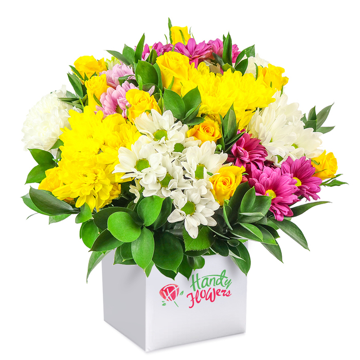 A lush, exuberant bouquet of mixed blooms rises from a pristine white square gift box, photographed against a stark white background that allows every colour to glow distinctly, as if lit by soft daylight drifting over New Barnet rooftops. The arrangement has a rounded, dome-like silhouette, densely filled so that no gaps of space are visible between the flowers. At its heart, generous clusters of yellow blossoms capture attention: full-headed yellow chrysanthemums with countless layered petals form rich, textured cushions, while smaller sprays of yellow roses, many still in tight bud, gather in golden clusters that suggest both abundance and new beginnings. Interlaced among these are numerous white flowers that brighten and cool the palette. Small daisy-like chrysanthemums, each with a neat corona of white petals around a clear green centre, dot the bouquet like little stars. Larger white chrysanthemums add body, their soft, slightly tousled petals overlapping to create a plush, almost velvety surface. Threaded throughout the design, vivid fuchsia and magenta chrysanthemums offer an eye-catching counterpoint, their saturated tones adding sophistication and drama, similar to the surprise of finding a hidden garden near Victoria Park. Some of these blooms show subtle hints of paler pink towards the tips, bringing gentle variation to the colour story. The whole composition is framed by glossy, dark green foliage that peeks between flowers and spills slightly over the edge of the box, its smooth, resilient leaves reinforcing the freshness of the arrangement. The contemporary white cube serves as a minimalist base, giving the piece a smart, gift-ready character suited to birthdays, get-well messages, or thank-you gestures for homes and workplaces around New Barnet. The bouquet feels joyful yet polished, like a thoughtfully chosen present ready for local delivery.