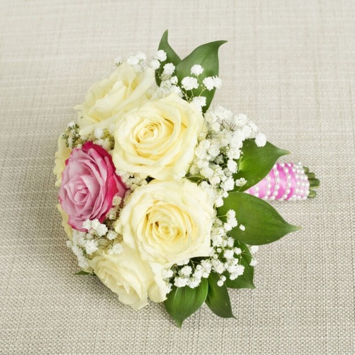 Viewed slightly from above, a compact and flawless bouquet is showcased atop an understated, beige linen background-calling to mind the timeless charm of New Barnet's historic boutiques. At its heart are five or six sumptuous cream or pale yellow roses, each bud fresh and tightly coiled, symbolising new love just beginning to unfold. A vivid fuchsia-pink rose peeks boldly from among the softer blooms, providing an irresistible focal point that echoes the vibrancy found in the colourful window displays of New Barnet's High Road shops. Generous clusters of airy white gypsophila thread their way throughout, lending a light, almost ethereal texture that contrasts beautifully with the voluptuous roses. Framing the bouquet are deep green leaves, glossy and abundant, suggesting both vitality and classic grace. The handle, crafted for comfort and style, is wrapped in bright fuchsia-pink satin ribbon, encircled by shining pearl strands wound in an elegant spiral. This detail feels at home in New Barnet, where tradition meets modern taste in every celebration. The ensemble rests against its lightly textured ground, the whole image radiating the sophisticated artistry and gentle warmth ideal for weddings, engagement parties, or sophisticated gatherings across North London-each event elevated by the thoughtful expertise and community spirit of local New Barnet florists.