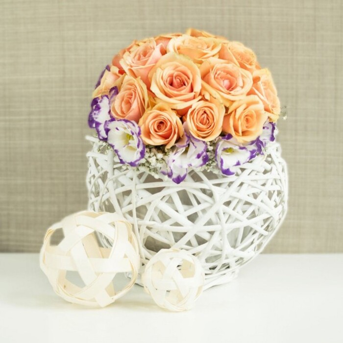 A sophisticated floral centerpiece is displayed against a softly textured beige background, like the gentle tones of a sunlit New Barnet townhouse wall. At the centre of the composition, a tightly gathered dome of peach and apricot roses rises from a low, rounded base, forming an almost perfectly spherical mound of petals. The roses are in full bloom, their layered petals unfolding in plush spirals that catch the light, moving from pale peach on the outer edges to richer apricot and soft salmon tones toward the centre. Encircling the lower edge of this rose globe is a collar of white lisianthus, each bloom edged with a dramatic band of deep violet or purple. The finely frilled petals create a lace-like effect, standing out crisply against both the warm roses above and the white container below. Tiny sprigs of white baby's breath are woven through this ring, their cloud-like clusters adding delicacy and filling any slight gaps with airy texture. The flowers are arranged in a distinctive, bulbous white container woven from rattan or wicker, its open, basket-like lattice allowing glimmers of the background to show through. This handcrafted vessel sits on a smooth white surface, its clean lines and bright tone adding a contemporary contrast to the natural weave. To one side, two decorative spheres made from pale, wood-toned strips echo the open, interlaced pattern of the vase-one larger, one smaller, positioned casually yet deliberately to enhance balance. The overall feel is one of refined calm and natural elegance, perfect as a wedding table arrangement for a reception near New Barnet station or a stylish dinner party overlooking the greenery around Oakhill Park, where the flowers bring a sense of fresh, tailored beauty indoors.