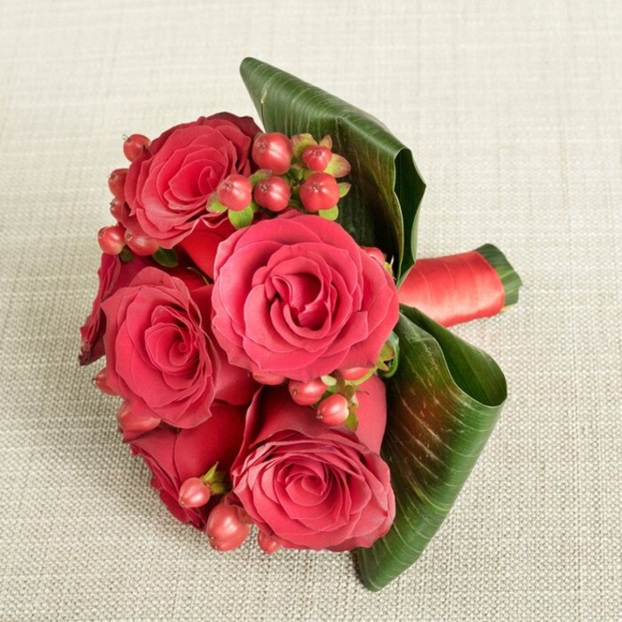A compact, hand-tied bouquet of deep red roses and berries lies gracefully on a light beige, finely textured fabric, the neutral background chosen to enhance the intensity of the blooms' colour. Around six roses dominate the arrangement, each one in full, luxurious bloom, their petals thick and velvety, curling outwards in layered spirals of crimson and wine-red. Between the roses, clusters of small Hypericum berries appear in rich shades of reddish-pink and deep red, the rounded forms adding a lively counterpoint to the soft, ruffled petals and creating a sense of rhythm throughout the design. Wide, dark green leaves have been wrapped and curved with an expert hand, forming an elegant frame around the right side of the bouquet and lending structure and depth, their subtle shine catching the gentle light. At the base, a rich red satin ribbon secures the stems, its smooth, slightly reflective surface harmonising with the opulent feel of the roses and suggesting a bouquet ready to be carried to an evening celebration in New Barnet, perhaps headed toward a reception near High Barnet station or a cosy restaurant off the High Road. The light falls evenly, without dramatic shadow, revealing every texture-the grain of the fabric, the gloss of the berries, the softness of each petal. It feels like a thoughtfully designed piece from Flowers New Barnet, ideal for an anniversary, proposal, or wedding party, evoking the warmth of shared moments and the classic romance that red roses bring to homes and venues across this peaceful corner of North London.
