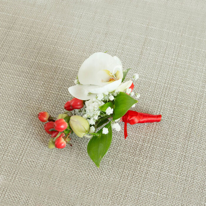 A finely detailed boutonniere or small corsage is photographed from a slightly elevated, top-down angle, resting on a light beige, woven fabric that resembles natural linen or burlap, evoking the relaxed yet polished style often chosen for weddings around New Barnet and the surrounding green spaces. At the centre of the composition is a pristine white Phalaenopsis orchid flower, its wide, gently arched petals appearing almost luminous against the neutral background. The orchid's throat carries a soft infusion of creamy yellow merging into subtle orange, creating a warm focal point that draws the eye inward. Crowning and framing this orchid are generous tufts of white baby's breath, each delicate floret like a tiny star, forming a cloudlike halo that contrasts in both scale and texture with the larger, smooth petals. Positioned just below and to one side is a small cluster of round, glossy red berries, their surface catching the light and providing a rich, festive accent that feels perfect for winter weddings or Christmas parties near Monken Hadley or Oakleigh Park. Interspersed with these are a handful of paler green, oval buds, hinting at berries still ripening and adding a sense of natural progression to the design. Surrounding the flowers and fruit, a selection of lush green foliage-some leaves broader and tapering to a point, others more slender and elongated-adds depth, framing the orchid and giving the arrangement a grounded, botanical character. The stems are carefully bound at the base with a vivid red satin ribbon, wrapped smoothly and tied in a compact knot, its short ends lying neatly along the fabric. This precise finishing touch suggests the piece is freshly prepared and ready to be pinned on a groom's lapel, worn by a prom guest, or used as an elegant corsage, the sort of handcrafted detail a local New Barnet florist might send out for same-day delivery before a special evening.