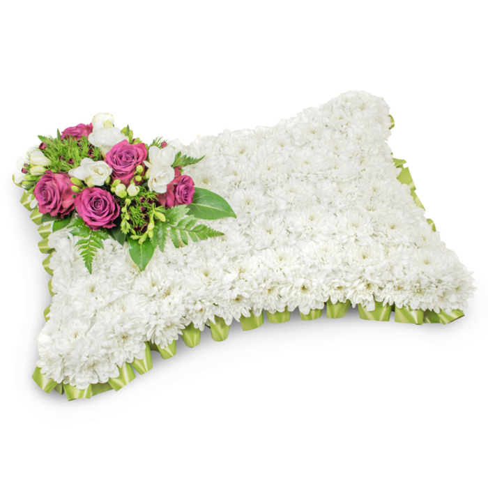 A classic pillow-shaped sympathy arrangement, expertly layered from hundreds of small white chrysanthemums to form a plush, uniform surface that reads like a gentle, floral cushion-soft in texture and serene in tone. The rectangular form, with softly rounded corners, is interrupted only by an elegant accent at the upper-left: a compact spray of deep magenta and fuchsia roses whose velvety petals open in measured layers, suggesting warmth and lasting affection. Interspersed among those roses are clusters of pure white freesias or similar tiny blooms, adding lightness, a hint of fragrance, and a luminous contrast to the strong rose colouring. Lush greenery-slender fern fronds and glossy foliage-fans beneath the spray and frames it naturally against the pure white expanse, offering depth and a sense of living support. A pleated, pale green satin ribbon trims the edges, its neat loops peeking from beneath the chrysanthemums to lend a subtle ceremonial finish and a soft ribboned outline. Set against a stark white backdrop with a gentle shadow, the pillow's three-dimensional form is clear and dignified. Intended as a funeral or graveside tribute, this arrangement communicates peace, eternal rest, and thoughtful remembrance. As a local New Barnet florist who often prepares tributes for services near Oakleigh Park and the Victoria Recreation Ground, I craft this design to be both comforting to touch in memory and quietly eloquent in presence-an arrangement that honours loss with refined simplicity and heartfelt care.