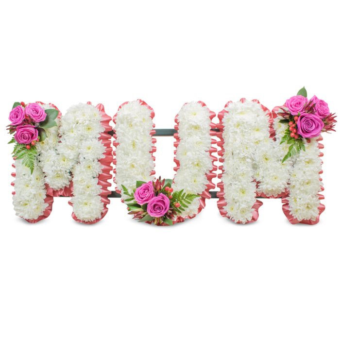 A refined and heartfelt memorial display spelling out MUM in three connected capital letters, photographed against a neutral white background to emphasize its clean lines and floral detail. Each letter's face is densely covered with small, pure white chrysanthemums that create a uniform, textured surface - the kind of soft, matte finish that reads as both formal and tender. The perimeter of each letter is softly lined with a ruffled pale-pink trim, a fabric or ribbon that introduces a gentle blush and a tactile, almost comforting edge to the otherwise monochrome letters. A subtle dark green support frame links the letters so the piece can stand steadily at a service or beside a graveside; the mechanics are discreet but dependable. Artistic clusters of deep fuchsia roses, fern-like greenery and tiny reddish-orange hypericum berries are positioned as finishing touches: on the upper left arc of the first M, in the lowest curve of the U, and on the upper right of the second M, giving three bright focal points that harmonise with the soft pink edging. The overall impression is one of considered craftsmanship - the crisp chrysanthemums suggest remembrance and purity, while the pink roses and ribbon whisper of affection and warmth. Ideal for a funeral service, wake or graveside ceremony in and around New Barnet - whether at a local church near Station Road or a quiet cemetery close to Brunswick Park - this tribute offers families a composed, comforting way to honour a beloved mother.