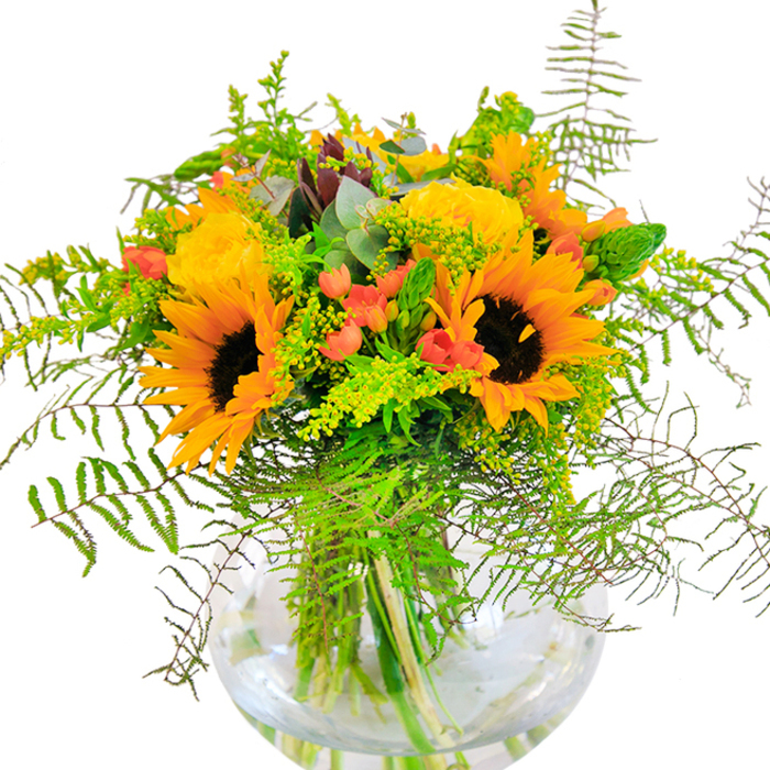 At first glance, this floral creation transports you to the sunniest corners of New Barnet, where vibrant blooms are artfully gathered by passionate florists who know the local rhythm. Gleaming sunflowers bloom confidently, their wide, amber petals and deep centers capturing the impression of uninterrupted sunlight. Twined with them, garden roses in buttery yellow slowly awaken, layers folding out in classic spirals, their fragrance mingling softly with the bouquet's greenery. Red Hypericum berries offer a dramatic punctuation, their glossy finish glinting among the gently arching ferns and filler, reminiscent of wild hedgerows seen along Mays Lane. Touches of goldenrod and slender green spikes weave through the design, adding finesse and texture, while eucalyptus leaves suggest calming shade beneath mature trees in Monken Hadley. The arrangement is cradled in a flawless, round glass vase that both magnifies the vivid stems and gives a contemporary clarity to the whole composition. Every detail-the velvet of a sunflower, the hint of citrus scent from the roses, the crisp freshness of intricate foliage-contributes to a bouquet that feels alive with the spirit of New Barnet. This joyful collection is perfect for birthdays, anniversaries, or simply sharing a sense of celebration and heartfelt warmth within the local community and beyond.