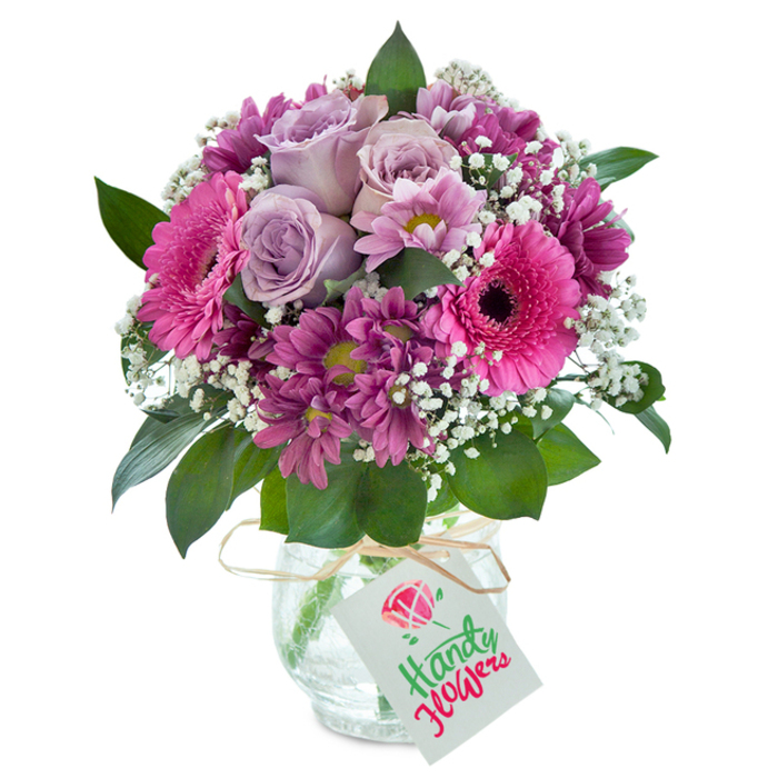 Centered in a clear, artfully textured glass vase, this lush arrangement offers a masterful composition of pink, purple, and white blooms, glowing softly under diffuse studio lighting. The visual heart of the bouquet is made up of opulent lavender roses, their petals opening in a tender spiral, surrounded by a lively ring of deep magenta chrysanthemums and bold fuchsia Gerbera daisies, each with a velvety dark centre. Interspersed among the main flowers, clouds of white baby's breath create a sense of lightness and grace, playing beautifully against the vivid colours and invoking memories of peaceful afternoons in New Barnet's green spaces. Lush, dark green foliage peeks through, framing the arrangement with an organic elegance reminiscent of the hedgerows lining the nearby parks. The vase itself, featuring a crackled finish, catches and refracts the light, adding an extra dimension of rustic charm. A simple tie of natural raffia hugs the vase just below the lip, from which a rectangular white gift tag dangles-its blank surface an invitation to personalize with a message for any special occasion, such as a birthday celebrated near Barnet High Street or a gesture of thanks for a friend living in the local community. Every detail, from the petal layering to textural interplay, showcases skilled New Barnet floristry-crafted for freshness, beauty, and heartfelt giving, ready for prompt delivery to homes, offices, and celebrations across the neighbourhood.