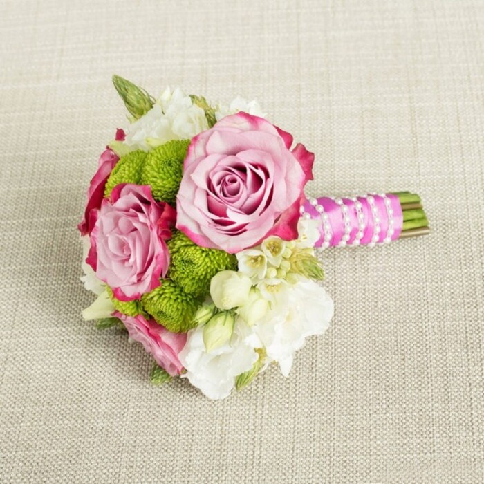 Set against a softly woven, light beige fabric backdrop that suggests a natural linen or burlap runner laid across a New Barnet reception table, a small hand-tied bouquet rests horizontally, its stems extending neatly to the right. The focal point of the arrangement is a trio of pink roses, two fully open and one slightly less mature, each bloom showcasing a graceful ombré transition from a pale, almost blush centre to deeper fuchsia and magenta tones at the gently ruffled outer petals. Their layered spirals draw the eye inward, evoking a sense of classic romance. Surrounding these roses are compact domes of vivid lime-green button chrysanthemums, their densely packed mini petals forming rounded, cushion-like shapes that lend texture and a fresh, contemporary twist to the palette. Interwoven throughout are creamy white filler flowers, perhaps delicate lisianthus or freesia, their soft, multi-petalled heads relaxing into the spaces between the bolder blooms to create a light, airy feel. Tiny, star-like white flowers with pinprick yellow centres add an extra dusting of detail, reminiscent of wax flowers often used in bridal work. Minimal dark green foliage peeks through, offering just enough contrast to ground the brighter shades. The stems are bound into a compact handle with a bright, fuchsia satin ribbon, the fabric smooth and slightly glossy in the light. Winding over this ribbon is a narrow strand of white faux pearls, carefully twisted into a diagonal pattern that gives the bouquet a gently glamorous, bridal character. This elegant, intimate arrangement, crafted in the style of Flowers New Barnet and suitable for bridesmaids, petite bridal posies, or small centerpieces at local venues around High Barnet and Hadley Common, radiates freshness, tenderness, and understated sophistication.