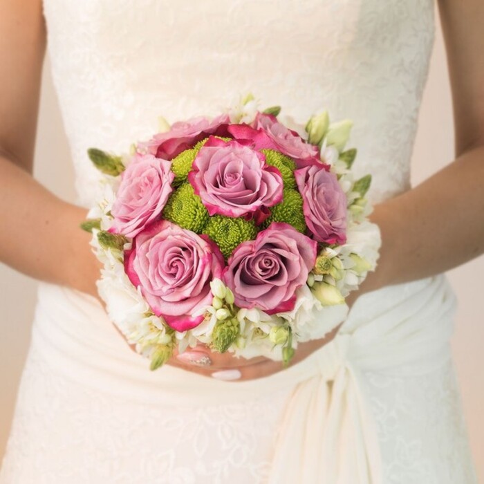 A striking, close-up portrait from Flowers New Barnet showcases a bride in the heart of New Barnet, her elegant lace gown providing a romantic backdrop as she holds a meticulously crafted, round bouquet. The bouquet is a celebration of both traditional wedding beauty and contemporary colour. At its centre, generously bloomed roses transition from the softest lavender at their core to more vivid, fuchsia-tipped petals, forming a lush, multidimensional crown. Around these, vivid lime-green button chrysanthemums-chosen for their joyful, modern texture-impart a lively twist, echoing the fresh energy of local florists near New Barnet railway or bustling High Road flower stalls. The arrangement is edged by a soft spray of crisp white blooms, possibly freesia or miniature roses, with slender green buds peeking through, adding depth and a breath of spring. The bride's hands are gently positioned, her pale pink nails tipped with a dash of sparkle, securing the bouquet as if holding a promise. Soft, even daylight illuminates the floral textures and the intricate patterns of her dress, while subtle folds in the gown's draping catch the light, highlighting artisan craftsmanship. This image encapsulates the thoughtful planning, joy, and detailed skill that define a New Barnet wedding florist's work-invoking the gentle romance, grace, and memorable palette perfect for a local bride's special day.