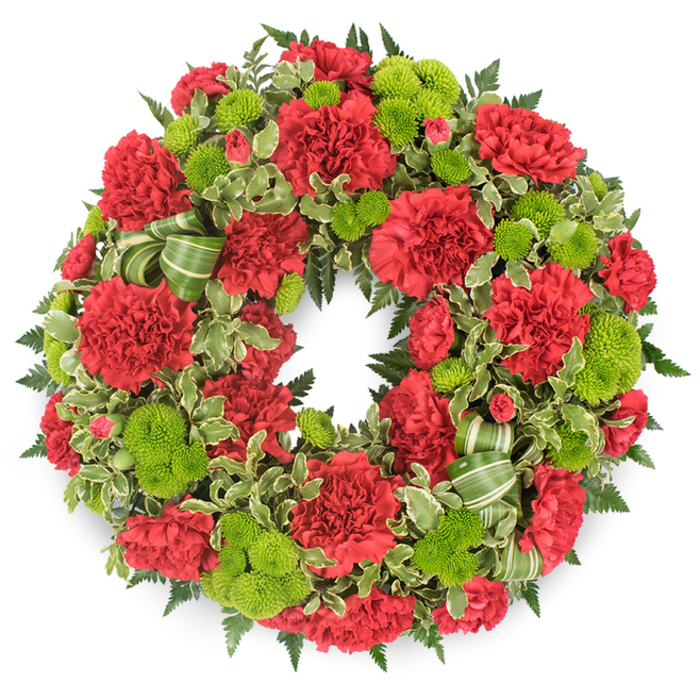 A traditional circular funeral wreath, rich with deep red carnations and zesty lime green button chrysanthemums, is shown in full focus against a stark white background that allows every nuance of the flowers and foliage to stand out. The ring is dense and luxurious, with no visible gaps, giving a sense of generous care and thoughtful craftsmanship. Red carnations dominate the design, their velvety, layered petals ruffling outwards to create soft cushions of colour that vary slightly in size and openness, mimicking the natural variation seen in a freshly picked garden. Threaded through this crimson field are rounded clusters of lime green pompon mums, their small, perfectly formed heads creating bright, lively dots of colour that illuminate the wreath and symbolise new life and hope. Beneath and around the flowers, a structured bed of greenery adds texture and depth: dark leatherleaf fern forms a shadowy, fern-feathered halo, while sprigs of variegated pittosporum bring lighter green tones and creamy white edges that frame the bolder blooms. A decorative ribbon in complementary greens is woven tastefully into the lower section, folded into gentle loops that echo the circular form and lend a subtle sense of movement. The composition feels serene and respectful, like something chosen for a funeral or memorial service in New Barnet, perhaps for a service near Barnet High Street or a quiet farewell close to Oakhill Park. Crafted with the sensitivity of an experienced local florist, the wreath conjures feelings of remembrance, love, and the wish to honour someone's life with beauty and dignity.
