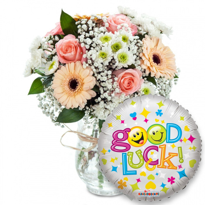 Lovingly arranged and photographed against a pristine white background, this 'Good Luck' gift pairs a textured floral bouquet with a festive foil balloon - a composition I would happily prepare at Flowers New Barnet for someone heading into an important new chapter. The bouquet fills a clear glass vase tied with rustic twine: powdery light pink roses with tight, velvety petals bring a gentle perfume, while peach-orange Gerbera daisies with deep brown centres read like cheerful, sunlit faces. Wispy sprays of Gypsophila (baby's breath) thread softly through the blooms, lending a lace-like delicacy, and tight clusters of small white-and-lime-green chrysanthemum florets inject a crisp, energising contrast. Dark green foliage peeks through the arrangement, grounding the pastel palette with natural depth. Dominating the right foreground, a round metallic silver balloon catches the light, its surface peppered with cheerful blue and pink hearts, yellow and blue stars, and tiny orange and green marks; Good Luck! arches across it in playful, multi-hued letters. The word Good is presented in bright purple with the two 'o's substituted by animated yellow smiley faces - one winks, the other sticks its tongue out - while Luck progresses in green, orange, red and blue, capped by a green exclamation. A small KALEIDOSCOPE logo appears at the balloon's lower edge. The overall mood is encouraging and handsomely balanced, ideal for exam days, job interviews, graduations or send-offs, and suited for local delivery within New Barnet - whether you're near Station Road, Ballards Lane or the community around Brunswick Park.