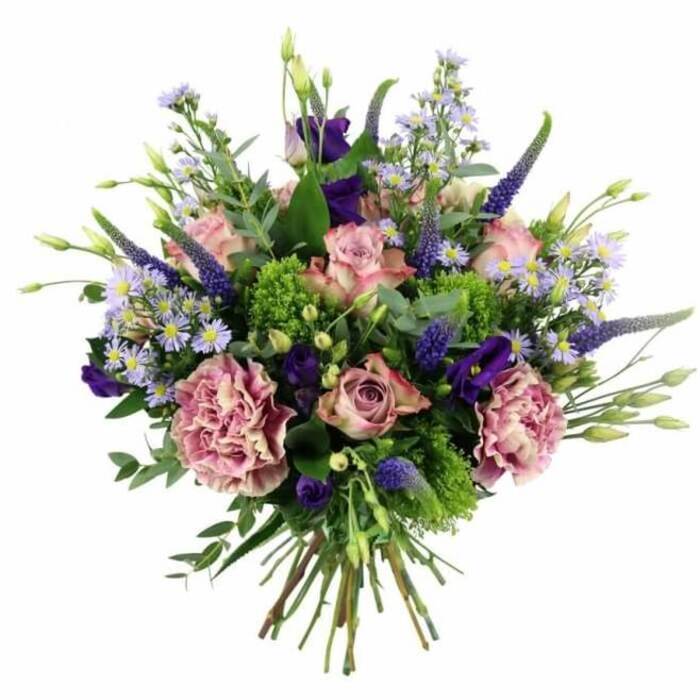 Set against a stark white backdrop, this hand-tied bouquet appears as if floating, its soft pinks, mauves, and purples forming a rounded, abundantly textured dome of colour. At its centre and scattered throughout are pastel pink roses, some still coyly cupped, others more fully opened so that the intricate spirals of their petals are clearly visible. A few blossoms carry a slightly deeper pink blush along their edges, giving a gentle, natural variation as you would expect from a skilled florist working with the finest stems. Embracing the roses are large, ruffled carnations in dusky rose and mauve tones, their layered frills lending a plush, almost fabric-like texture that fills any gaps and makes the bouquet feel generous and full. Punctuating this softness, tall Veronica stems rise in deep lavender-blue, slender and slightly curved, adding vertical movement that leads the eye from the tightly gathered stems to the highest flower tips. Nestled among them, rich purple lisianthus blooms appear like small bells, some open and some in bud, intensifying the colour story and adding depth. Delicate sprays of star-shaped, aster-like flowers in pale lavender-blue weave through the bouquet, giving lightness and a feathery, garden-inspired charm. The flowers are cradled in a collar of mixed foliage, where fresh lime-green filler contrasts with muted eucalyptus and other greenery, echoing the fresh, leafy feel of New Barnet's residential streets and nearby green spaces. The neatly tied stems at the base confirm this is a classic hand-tied design, ideal for same-day delivery to homes and offices around New Barnet, perhaps placed in a hallway to greet guests or on a dining table for a birthday, anniversary, or intimate family gathering.