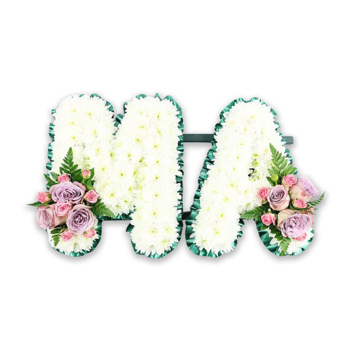This photograph shows a refined letter tribute spelling MA in large, block capitals, styled as a funeral or memorial arrangement and presented against a plain white backdrop that emphasises its structure. The letters are generously filled with pure white chrysanthemums, each stem placed close to the next so no gaps appear, creating a thick, luxurious bed of petals. The chrysanthemums have that familiar, slightly feathery texture, catching the light and giving the letters a gentle, cloud-like softness despite their firm shape. Encircling both the M and the A is a deep green ribbon border, likely satin, folded into neat pleats so that the edge looks both decorative and precise. It adds contrast and definition, making the white centres stand out boldly as you might see them laid at a local New Barnet chapel or crematorium. At the lower left corner of the M and the lower right of the A, elegant accent sprays soften the tribute with pastel colour. Each spray features open lilac roses, pale pink roses, and smaller pink spray roses, layered together to create gentle depth. A few fern fronds and clusters of rich green foliage weave through these roses, bringing an airy, natural feel that echoes the greenery of New Barnet's residential gardens and tree-lined streets. The overall arrangement has a balanced, symmetrical composition, suggesting careful thought and professional skill by a florist familiar with creating personal tributes for local families. The piece clearly honours someone known lovingly as Ma or Mama, combining formal white sympathy flowers with tender pink and purple hues to express enduring love, respect, and a peaceful farewell.