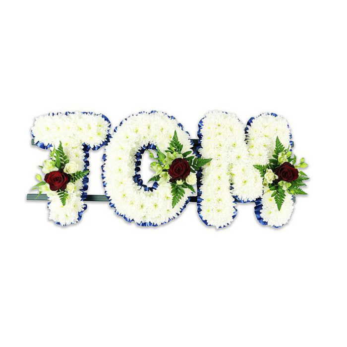 This personalized floral tribute forms the name TOM in voluminous, sculpted letters entirely sheathed in small, pristine white chrysanthemums, producing a soft, lamp-like luminosity across each surface. The letters' perimeters are neatly finished with a vibrant royal blue ribbon that provides crisp contrast and formal definition, allowing the white blooms to stand out against a simple white background. Each letter carries a compact floral accent arranged with classical restraint: a single, intense dark red rose anchors the spray, its velvety curves speaking of affection and sorrow, while tiny ivory spray roses and sprigs of lush green ferns and other foliage nestle beside it, offering texture, tonal variation, and a sense of gentle movement. A subtle dark green or black metal bar runs beneath the composition, visible only at the base, indicating the professional support system that keeps the tribute upright during a service. The overall mood is one of composed reverence-the chrysanthemum clusters feel tender to the touch, the rose offers a quiet scent, and the blue ribbon frames the message with dignity. As a design commonly requested for funerals and memorials around New Barnet and nearby Monken Hadley Common, it's the kind of handcrafted, bespoke lettering our local florists make for families seeking a meaningful, elegant farewell that reads clearly in both intimate gatherings and photographed memorials.