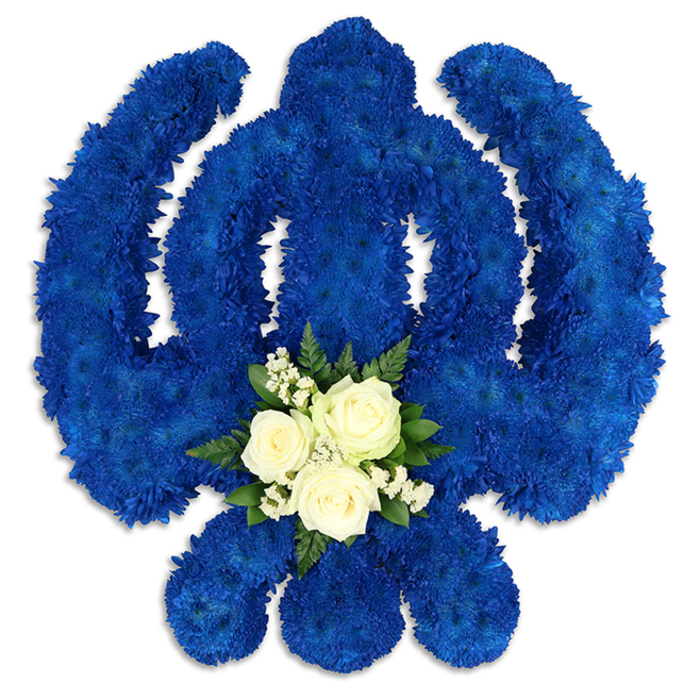 This image captures an elaborate Sikh Khanda floral tribute, carefully arranged to create a bold yet reverent centrepiece, perfect for a Sikh funeral or memorial service in New Barnet. The sacred Khanda emblem is precisely formed: a strong central sword rising vertically, a smooth circular chakkar encircling it, and two symmetrical kirpans sweeping outward on either side, their curved silhouettes clearly defined. Every part of the symbol is constructed from countless royal blue chrysanthemum florets or similar small-headed blooms, packed closely together so no gaps break the intensity of colour. The effect is a luxurious, velvety surface, with the deep blue shade invoking themes of loyalty, steadfast faith, and spiritual depth. Set against a bright white background, the edges of the Khanda appear crisp and exact, while gentle shadows around the petals introduce a subtle three-dimensional feel, as if the design has been raised slightly from the surface. At the lower part of the central blade, a contrasting focal point draws the eye inward. Three immaculate white roses, with neatly layered petals, are nestled within a cloud of delicate white filler flowers, likely baby's breath, and framed by small sprays of glossy dark green foliage. Together, these lighter elements soften the strong blue form, suggesting peace, purity of intention, and a blessing for the soul of the deceased. The arrangement feels both modern and deeply traditional, echoing the cared-for streets and diverse community of New Barnet, where families often seek bespoke tributes that honour faith and culture. This Khanda tribute speaks of a florist's patience and understanding, the kind of careful work one might commission for a solemn service or crematorium farewell nearby.