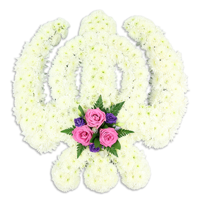 A striking Sikh Khanda floral tribute dominates the image, its sacred geometry rendered with meticulous precision in snowy white chrysanthemums. The central double-edged sword rises upright, framed perfectly by the circular chakkar, while the two kirpans curve protectively at the base, their outlines crisp and unmistakable despite being formed entirely from petals. Each chrysanthemum head is tightly nestled against the next, creating a dense, velvety surface with subtle shadows between the blooms, giving the whole piece a sculpted, almost carved appearance. The white of the flowers feels soft rather than harsh, a gentle ivory that recalls the quiet light of an overcast morning in New Barnet. At the lower centre of the Khanda, just where the kirpans meet, a vibrant posy of accent flowers introduces warmth and tenderness. Three medium-sized pink roses take pride of place, their petals layered like delicate silk, some slightly cupped, others beginning to open more fully. They are framed by smaller, deep purple blooms, likely lisianthus or freesias, which add depth and a touch of solemn richness. Fine sprays of dark green fern and other foliage peek between the flowers, giving texture and a natural, garden-fresh feel. There are no visible ribbons or text; the tribute speaks purely through form and colour. Set against the plain white backdrop, a faint shadow beneath the arrangement hints at its three-dimensional depth, as if it could be resting on a stand in a quiet chapel or family home near New Barnet. This floral Khanda feels both powerful and gentle, an eloquent expression of Sikh faith, remembrance, and respect at a funeral or memorial service.