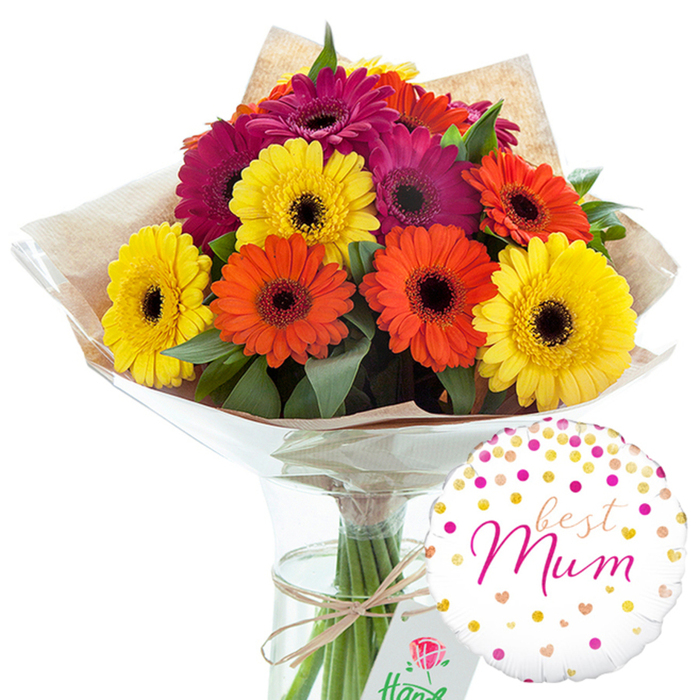 A thoughtfully arranged Mother's Day-style gift is captured against a clean studio backdrop, featuring a generous bouquet of gerbera daisies in a glass vase, alongside a cheerful best Mum balloon. The bouquet is full and rounded, with around a dozen large gerbera blooms positioned closely together so their colour blocks blend harmoniously. Petals in sunny yellow, fiery orange, and saturated magenta fan out evenly from dark central disks, creating a striking contrast that draws immediate attention. The colours feel warm and energetic, reminiscent of late afternoon light over New Barnet's tree-lined streets. Between the flowers, rich green foliage peeks through, providing a fresh, leafy frame that softens the bright tones and adds visual depth. The stems are carefully hand-tied, then wrapped in textured, light brown kraft paper that looks rustic and slightly crinkled, suggesting a personal, handcrafted touch by a local florist. Over this sits a smooth sheath of clear cellophane, giving the arrangement a polished, gift-ready finish while still allowing the natural textures beneath to show. The bouquet stands upright in a tall, slender clear glass vase, filled with water that magnifies the parallel green stems inside. Around the neck of the vase, a simple bow of natural twine adds a final, understated accent. To the lower right, a round white foil balloon balances the composition. It is sprinkled with scattered gold, bronze, and bright pink polka dots and tiny hearts, framing the message best Mum in flowing pink script at its centre. The whole image conveys warmth, appreciation, and quiet celebration, ideal for a New Barnet family wishing to surprise a mother figure with a colourful, heartfelt floral delivery.