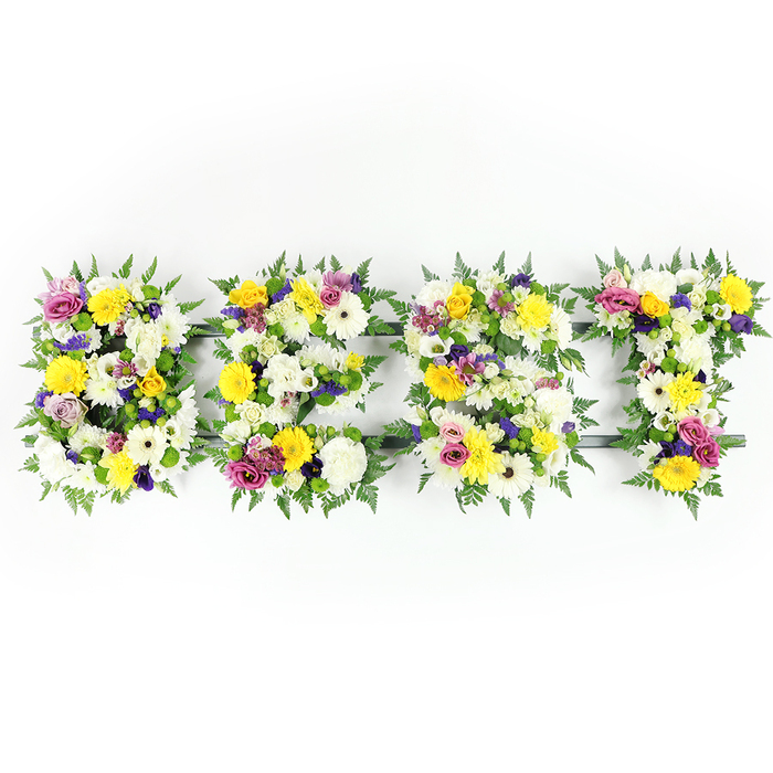 A lush, horizontal floral inscription spelling BEST is arranged in four substantial letters, each densely packed with fresh blooms and framed with rich green fern. The colour story is bright and consoling: pristine white chrysanthemums and creamy gerbera-like blooms sit alongside sunny yellow roses and gerberas, while soft purple lisianthus and deeper magenta-pink spray roses or carnations provide elegant, sympathetic contrast. Small clusters of pale green and white filler flowers nestle among the larger heads, giving the letters textured depth and a gently undulating surface that reads beautifully from a distance. Dark-green fern fronds curl around the perimeters, softening edges and adding a timeless, natural border.
Beneath the floral skin, a subtle dark green or metal framework supports the scale of the piece; you can glimpse this support through small gaps in the dense packing, particularly along the horizontal sections of the E and the crossbar of the T. The photograph is taken head-on with even, bright lighting, which clarifies petal structure, the satiny sheen of rose petals, and the feathery crispness of fern - and suggests the faint, comforting scent of roses and freshly cut foliage. This type of lettered arrangement is often selected for memorials, funerals, and commemorations to express love, admiration, or to celebrate a cherished life.
Flowers New Barnet made this tribute with local care, drawing on our experience delivering to New Barnet High Street, Brunswick Park, and nearby East Barnet Village; we craft lettering to read clearly in service settings and to provide a warm, handcrafted message of remembrance or celebration.