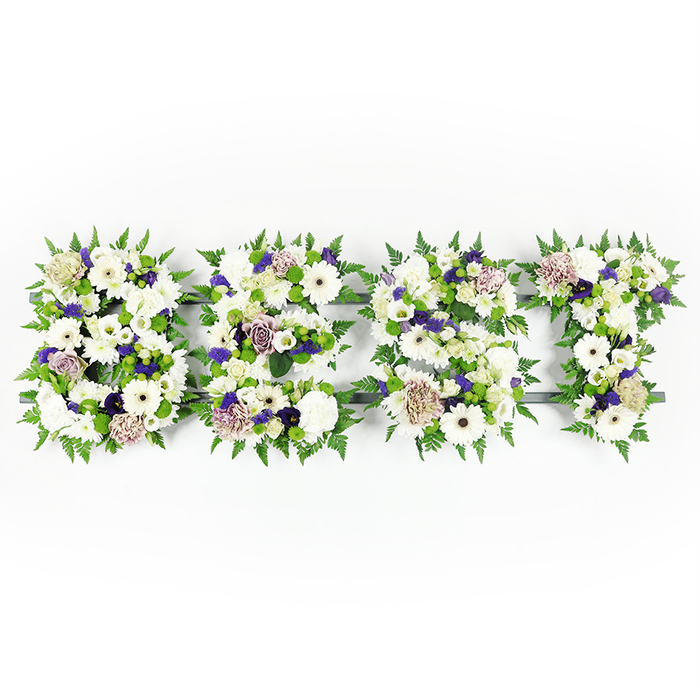 A horizontal tribute, carefully hand-built to spell the word BEST, occupies the frame in a calm, measured composition against a bright white background that makes every bloom read with delicate clarity. Each letter - B, E, S and T - is densely packed with pristine white flowers: large, immaculate Gerbera daisies with their dark centres, layered white lisianthus (eustoma) and full, cushiony carnations that together create a tapestry of soft, reflective whites. Soft mauve and lavender roses and ruffled purple carnations are woven through these whites, their muted purples lending a gentle dignity and depth that avoids gaudiness while suggesting reverence. Deeper purple filler flowers appear like thoughtful punctuation, small and deliberate, while compact green button chrysanthemums and sprigs of fresh foliage bring green vitality into the heart of each letter. The perimeter of each character is outlined with feathery fern fronds, whose soft, serrated edges cradle the blooms and shape the lettering with a natural, sympathetic contour. A slim, dark structural base gives the letters definition in places, visible only where petals lift, and it helps the tribute sit securely on a memorial stand. I imagine this design arriving at a chapel near Station Road or being placed at a service in the local area, where its quiet palette and careful construction offer a personalised expression of love and remembrance - perhaps reading as best loved or as a final, tender message from family and friends.