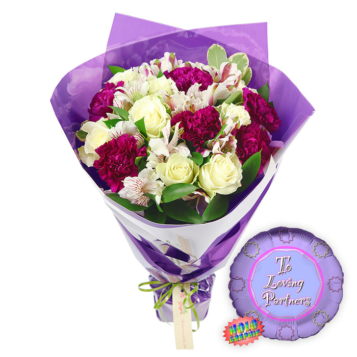 A carefully composed romantic gift set is arranged against a bright, uncluttered white background, drawing full attention to the interplay of flowers, colour, and light. The hand-tied bouquet at centre stage features approximately a dozen creamy white roses, their spiralled petals still closely held and velvety, exuding a sense of quiet luxury. Nestled among them are rich magenta and deep burgundy carnations, their densely frilled petals forming plush, cushiony blooms that bring depth and intensity. Fine white alstroemeria blooms weave through the arrangement, each slender petal etched with soft reddish-brown speckles and stripes, adding intricate patterning that recalls the delicate details a florist in New Barnet notices while working at the counter on a busy High Road afternoon. Around the flowers, layers of fresh greenery, including glossy green leaves and softly variegated foliage, unfold like a natural frame, offering contrast to the pale roses and jewel-toned carnations. The wrapping is thoughtfully styled in two layers: a crisp white paper underlayer, angled diagonally to support the stems, and an outer sheath of translucent purple cellophane that crinkles gently and rises around the heads of the flowers like a luminous veil. The base is cinched with a slim, bright green satin ribbon tied in a modest bow, from which a small, light brown tag is suspended, ready to bear a loving note. To the right of the bouquet rests a metallic purple foil balloon with a scalloped, medal-like edge, its centre a paler lavender circle inscribed in elegant pink script with the words To Loving Partners. A small rainbow HOLOGRAPHIC style sticker along the edge hints at a shimmering finish. This evocative pairing of flowers and balloon suggests an intimate romantic celebration, such as an anniversary or special date night, delivered to homes across New Barnet and nearby streets.
