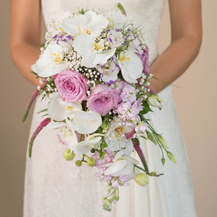 A sweeping close-up set against the understated tones of a New Barnet venue reveals a bride holding an opulent, cascading bouquet. The design is a showpiece of high floral artistry: central clusters of snow-white Phalaenopsis orchids, with their delicate, overlapping petals and golden-yellow throats, arch gracefully downward in a dramatic cascade. Complementing the orchids, velvety roses in pale pink and lilac shimmer in the gentle light, their fullness bespeaking love and celebration. Interlaced within are gossamer sprays of white baby's breath, lending both movement and the ethereal hint of a misty morning along East Barnet's tree-lined avenues. Subtle additions of mauve and lavender stock, along with festoons of fuchsia and magenta amaranthus, bring unexpected texture and vibrant colour. Touches of green foliage and slender, twisting buds provide natural detail, enhancing the bouquet's sense of freshness, as if just gathered from a New Barnet garden in high summer. The bride's gown-in ivory silk and lace-completes the scene, its intricate patterns echoing floral filigree. The result is a lush, romantic presentation, perfectly tailored for local weddings, anniversary celebrations, or anyone drawn to the refined beauty of expertly arranged flowers in the heart of New Barnet.