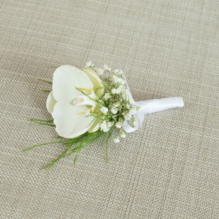 A refined white orchid boutonniere lies peacefully on a light tan, woven fabric surface, the texture of the material suggesting natural linen or soft burlap, chosen specifically to frame the flowers without competing with them. At its centre, a flawless Phalaenopsis orchid bloom opens in wide, sculpted petals, the white so clean it appears almost luminescent against the muted background. The petals curl gently, guiding attention inward to a throat tinged with soft yellow and cream, adding a whisper of colour that suggests vitality and freshness, as if the flower has just been prepared in a New Barnet florist's studio minutes before a ceremony. Surrounding this central orchid is a halo of baby's breath, their countless tiny white blossoms gathering in airy clusters, each no larger than a pinhead, yet collectively forming a soft, cloud-like border. Between these small flowers weave slender stems of bright green foliage, possibly maidenhair or asparagus fern, bringing a touch of woodland grace and offering a fresh, slightly wild counterpoint to the orchid's poised formality. The stems of the orchid, gypsophila, and greenery are pulled neatly together and wrapped with a strip of matte white satin ribbon, the wrapping tight and smooth, ending in a discreet, well-formed bow that indicates careful, practiced hands at work, like those in a long-established New Barnet flower shop. Gentle, diffused light illuminates the scene, revealing the faint sheen of the orchid petals, the matte delicacy of the baby's breath, and the fine threads of the fabric beneath. The overall impression is one of timeless elegance and understated romance, perfect for a groom's lapel at a New Barnet wedding near local landmarks or for an intimate civil ceremony where simple, classic florals convey heartfelt emotion.
