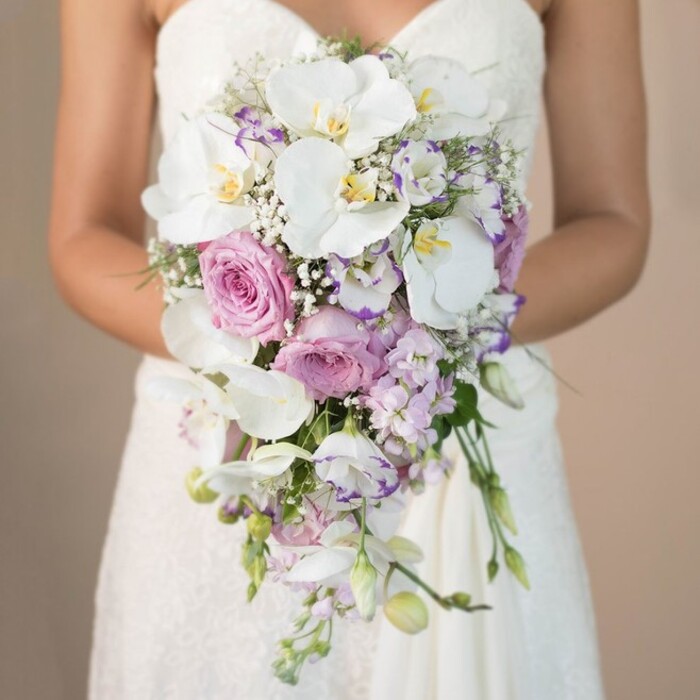 A luminous wedding scene unfolds as a bride, dressed in a finely tailored strapless white gown featuring delicate lace motifs, poses in a softly diffused beige setting suggestive of tranquil New Barnet mornings. Her bouquet, crafted in the classic trailing teardrop shape favoured by many local couples, is an arresting focal point. Wide, flawless white phalaenopsis orchids with lemon-yellow centres anchor the design, their broad, lustrous petals imparting a sense of luxury and sophistication. Amidst the orchids, generous clusters of pastel pink garden roses bloom with velvety richness, each petal curled as though shaped by the gentle touch of a New Barnet florist. Wispy lavender and light purple stocks thread through the composition, their upright forms introducing softness and subtle texture. Interlaced white lisianthus, some petals blushed in violet, add another layer of charm and delicacy. Feathery bunches of baby's breath confer a light-as-air fullness, their snow-white florets glowing in the ambient light. Sparse sprigs of greenery, tastefully woven between floral highlights, maintain a restrained balance and reinforce the bouquet's organic beauty. The arrangement sweeps downward in a fluid cascade, mirroring the undulating lines of the bride's lace skirt as she stands serenely, perhaps dreaming of her walk down a New Barnet aisle. This image, rich with floral artistry and local character, perfectly illustrates the romantic, sophisticated style customarily prepared by neighbourhood florists for weddings and bridal events throughout New Barnet.