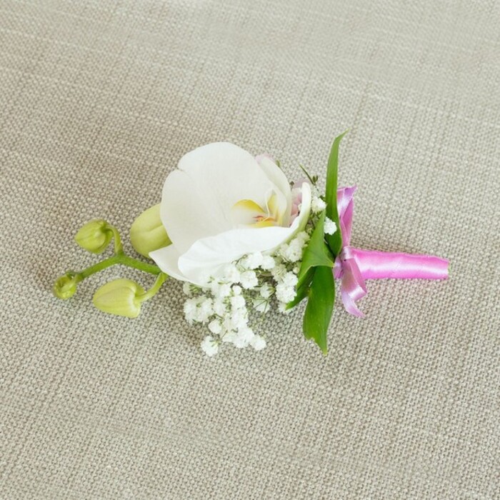 A refined orchid boutonniere is presented horizontally on a soft, light beige linen-like fabric, the subtle weave of the material creating a calm, neutral stage that feels very much like a thoughtfully set dressing table in a New Barnet townhouse. At the centre of this small floral composition lies a single, immaculate white Phalaenopsis orchid, its wide, velvety petals gently curving outward and creating a sense of openness and calm. The orchid's throat shows a tender wash of soft yellow with delicate touches of pale pink, adding just enough colour to draw the eye inward without disturbing the overall purity of the bloom. Encircling the base of the orchid, small sprays of white baby's breath form a delicate cushion, each tiny flower like a miniature star, adding volume and an airy, cloud-like softness. A few narrow, glossy green leaves peek out from behind the blossoms, their fresh, springlike hue bringing life and contrast to the design. To one side, a slender stem reaches out, bearing tightly closed, pale green orchid buds that hint at blossoms yet to unfurl, echoing the feeling of anticipation before a wedding or prom in local venues near New Barnet station. The stems are neatly gathered and wrapped with a slim band of bright fuchsia pink satin ribbon, tied into a tidy, elegant bow. The ribbon's sheen catches the light, introducing a playful yet sophisticated pop of colour that complements the cool white orchid. Gentle, even lighting highlights every texture-from the linen background and the satin bow to the smooth petals-giving the impression of a carefully crafted, timeless accessory ready for a groom's lapel or a special guest's outfit. Created with the attention to detail you'd expect from an experienced North London florist, this boutonniere radiates understated celebration.