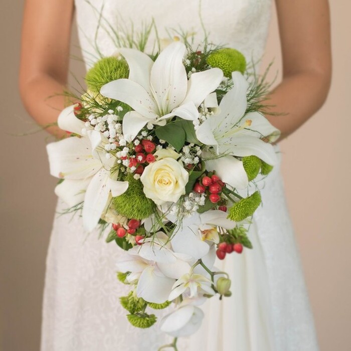 A bride in New Barnet is pictured at chest level, elegantly clutching a breathtaking cascading wedding bouquet against the softly blurred, ivory lace of her gown. The bouquet features an artful arrangement of snow-white lilies, each blossom broad and beautifully contoured, adorned with subtle russet speckles that bring depth to their radiant petals. At the center lies a perfectly shaped creamy rose, tightly curled and serene, anchoring the composition with a touch of perennial romance. Sprinkled throughout are vibrant green cushion chrysanthemums, their textured domes infusing energy and modernity reflective of New Barnet's thriving cultural scene. Accentuating the snowy blooms, festive red Hypericum berries burst between petals, offering visual warmth and lively contrast reminiscent of the area's seasonal festivities. Wispy clouds of baby's breath float across the bouquet, softening the scene with airy delicacy, while threads of fresh green foliage and slender ornamental grasses reach outwards to frame the arrangement organically. White Phalaenopsis orchids sweep graciously downwards, their trailing petals creating a dramatic waterfall effect, reminiscent of graceful lines found in local gardens and parks. The bride's hands, partially visible, hold the bouquet just above her gently patterned gown, uniting tactile detail with floral elegance. Bathed in gentle daylight, every texture and hue emerges vividly, providing a sense of hope, abundance, and refined celebration-a bouquet style that stands as a New Barnet favourite, sure to inspire couples dreaming of a timeless yet unique wedding statement.