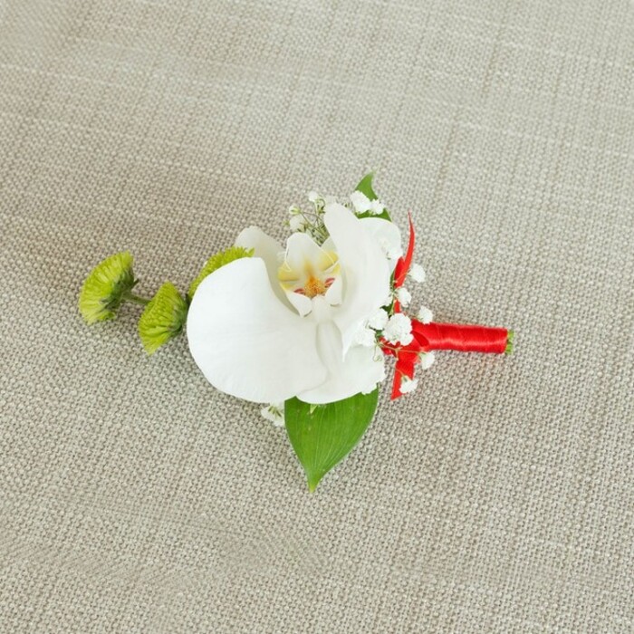 A sophisticated white orchid boutonniere is displayed on a softly textured, light beige linen surface, the neutral background allowing every floral detail to stand out with quiet confidence. At the core of the piece, a single Phalaenopsis orchid blossom draws the eye, its large, pure-white petals unfurling in a smooth, gentle arc reminiscent of folded silk. The centre of the orchid reveals a gentle wash of yellow that deepens into soft reddish-orange accents, lending warmth and complexity to the otherwise cool white bloom. Nestled closely around this focal flower are clusters of dainty white baby's breath, their countless tiny blossoms forming a delicate cloud that softens the silhouette and adds a romantic, almost ethereal texture. To the left side, two round lime-green chrysanthemum or green pom-pom flowers bring a lively pop of colour and a slightly spiky, intricate surface that contrasts with the orchid's sleekness, calling to mind the fresh, bright greens seen in gardens near Victoria Park in New Barnet. A broad, deep green leaf is tucked beneath the orchid, acting as a natural base that anchors the design, while a smaller, darker leaf appears near the bottom, adding visual balance. The stems are firmly bound by a vivid crimson satin ribbon, wrapped smoothly and finished with a compact bow or knot that introduces a confident, celebratory edge. The red ribbon stands out vividly against the beige fabric and white flowers, hinting at love, joy, and the formality of a wedding or black-tie event. Crafted with the attentive detail you'd expect from an experienced local florist, this boutonniere feels ideal for a groom or groomsman attending a New Barnet ceremony, ready to be pinned on just before the first photographs are taken.