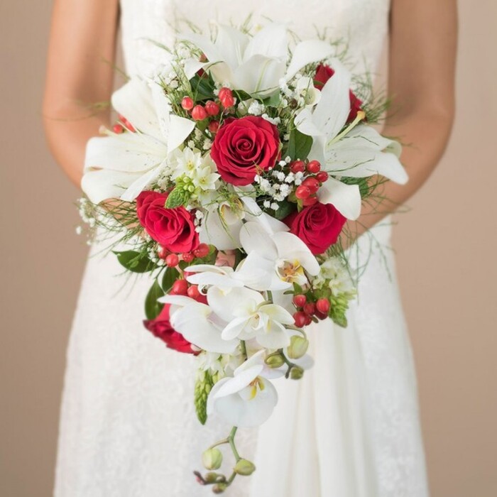 Central to this evocative New Barnet wedding scene is a stunning, teardrop-shaped bridal bouquet, showcased in the gentle glow of natural, diffused light. The composition features the bride, softly out of focus from the waist up, gently clasping the bouquet with both hands as her white lace gown offers a hint of classic elegance, the subtle detail of the fabric mirrored in the meticulous arrangement of blooms. Dominating the bouquet are magnificent white lilies, their broad petals curled open and contrasting beautifully against lush, full-bodied crimson roses-flowers that speak to tradition and enduring love, much like the community spirit found in New Barnet's historic lanes. Interspersed among the main blooms are graceful white orchids that spill downward, trailing with the finesse and harmony of a carefully composed musical phrase. Pops of colour emerge from vibrant red berries-reminiscent of festive wreaths or hedgerow finds in nearby parks-lending energy and playful texture. Airy sprigs of baby's breath and other tiny white blossoms interweave throughout, softening the overall effect and creating a romantic, cloudlike finish. The rich backdrop of feathery green foliage, featuring both wispy and spiky varieties, grounds the arrangement and provides dimension. The neutral beige background subtly guides all attention to the stunning interplay of tones and textures captured in this moment. It's a bouquet for those whose weddings blend classic beauty with modern flair-a perfect choice for couples saying their vows in New Barnet's elegant venues or charming gardens, crafted with heartfelt care by local floral artists experienced in making memorable occasions come to life.