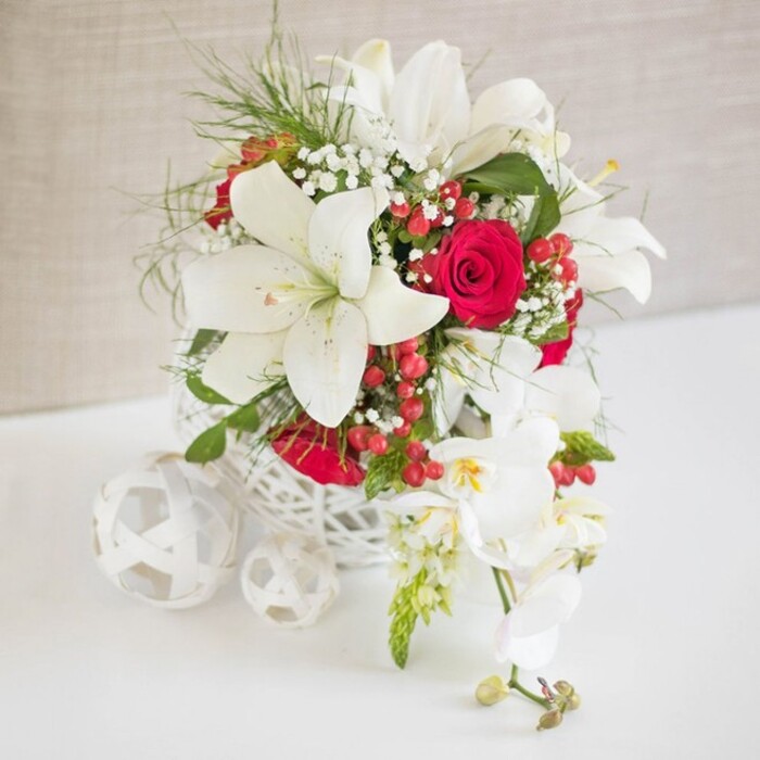 An artfully composed white and red floral arrangement stands centered in the frame, evoking the polished calm of a wedding breakfast laid out in one of New Barnet's elegant homes. The flowers are gathered in a round, basket-like container crafted from white open-weave strands, giving a sense of lightness and handcrafted charm as it rests on a spotless white tabletop. Emerging from this base, generous white oriental lilies unfurl their broad, six-petalled blooms, each flower showcasing long stamens tipped with earthy pollen, adding a subtle golden warmth amidst the cool whites. Nestled close to the lilies, sumptuous red roses in a deep, velvety shade bring a rich romantic contrast, their petals layered densely in full, rounded heads. Threaded between these statement blooms are delicate sprays of white baby's breath, creating a misty, cloud-like texture that softens the overall silhouette, while wisps of bright green foliage provide movement and a hint of freshly cut garden stems. Scattered throughout, polished red hypericum berries gleam like tiny ornaments, enhancing the festive palette and making the arrangement feel ideal for winter weddings, Christmas receptions, or special family gatherings in New Barnet and nearby Hadley. Flowing gracefully from the right side of the main composition, a cascade of pristine white Phalaenopsis orchids arcs downward, each flower perfectly formed with a hint of yellow and soft blush pink at the centre, accompanied by a few green-tipped buds. On the left-hand side of the scene, two or three white woven spheres, echoing the container's texture, are casually grouped on the table, adding a sculptural, modern touch. The backdrop, a light beige textured fabric, gently blurs into softness, ensuring the interplay of white, red, and green remains the clear focal point. The overall mood is one of sophisticated celebration, suitable for bridal table décor, anniversary dinners, or elegant seasonal entertaining across the EN4 area.