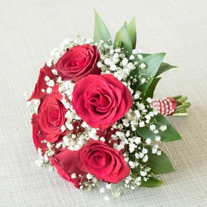 A lush, compact bouquet lies serenely atop a textured beige background, exuding classic romance through its careful arrangement of five to six fully opened, deep red roses. Each rose is a study in detail, with tightly furled centers giving way to layered, velvety petals that radiate a rich crimson glow, embodying timeless love. Between these signature blooms, an abundance of soft, white baby's breath-Gypsophila-spills outward, their cloud-like clusters brightening the bouquet and adding a delicate, airy feel, like the gentle morning fog drifting over Oakleigh Park. Fresh green leaves offer vivid contrast, their edges peeking around the flowers to add depth and a hint of the countryside surrounding New Barnet. The stems, cut and aligned with care, are expertly wrapped in a bold red satin ribbon, accentuated with a spiral of small, gleaming pearls for a subtle, elegant shimmer. The entire arrangement is presented against a neutral, softly textured surface that subtly elevates the intensity of the florals' colours and intricate details. Created by a Florist in New Barnet, this radiant bouquet is infused with artistry and heart, making it an ideal offering for weddings, anniversaries, or as a classic romantic gesture-its freshness and beauty enhanced by the expertise of the local floral community.