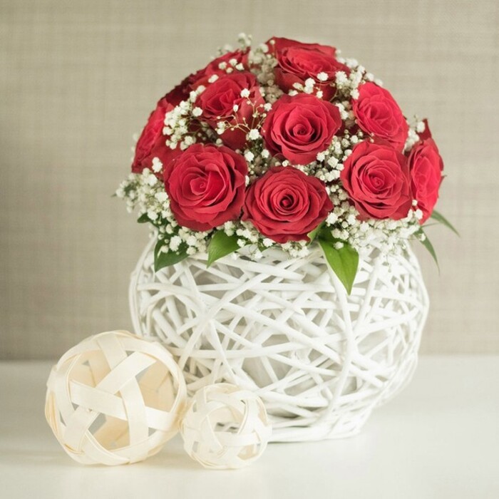 This image portrays an elegant floral centrepiece that could easily be imagined on a wedding table in New Barnet, its focal point a rounded cluster of deep red roses. Approximately a dozen or slightly more roses are packed closely together, forming a neat, spherical mound where each bloom is fully open, revealing rich, velvety petals that spiral inward in classic romantic fashion. The colour is a saturated crimson, intense yet softened by the presence of abundant white baby's breath threaded throughout the bouquet. The gypsophila forms delicate, airy sprays, each tiny blossom like a dot of light against the darker roses, adding contrast and a cloud-like softness that keeps the design from feeling too heavy. Here and there, small green leaves peek from between the flowers, introducing a whisper of natural foliage. Supporting this composition is a distinctive, rounded white vase with an open-weave structure. Thin, white, branch-like strands criss-cross and curl around one another, creating a hollow, basket-like globe that feels both contemporary and handcrafted, as if carefully woven by an artisan. The vase rests on a smooth, clean white surface that reflects the soft ambient light. To the left of the arrangement sit two decorative spheres in an off-white or pale beige tone, also made from flat, woven strips with an open lattice design; the larger ball is set slightly closer to the viewer, with the smaller one tucked behind, giving depth and a sense of relaxed styling. The backdrop is a gently textured, neutral wall in light beige, unobtrusive yet warm, like the interior of a calm New Barnet home along Station Road. The overall mood is refined, romantic, and perfectly suited to intimate celebrations, anniversary dinners, or wedding reception tables.