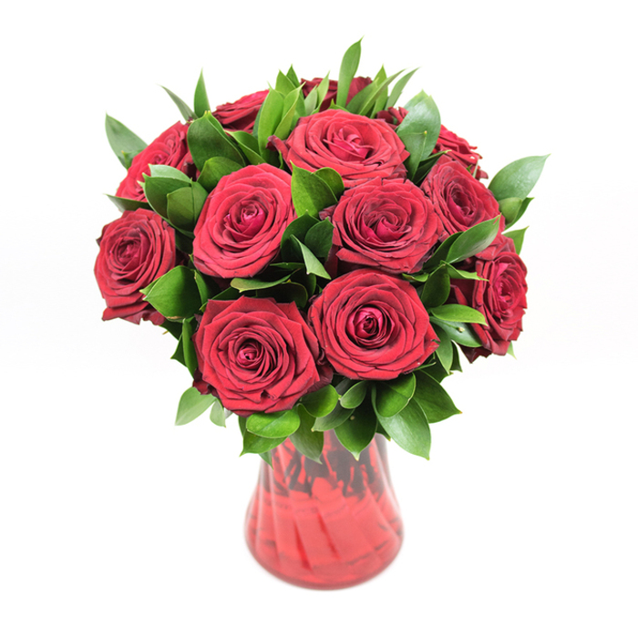 A richly coloured bouquet of deep crimson roses fills a translucent red glass vase, captured in front of a pristine white background that makes every hue and texture vividly clear. There are roughly twelve to fifteen roses, gathered into a classic, rounded formation, each bloom fully open but still neat, with tightly layered, velvety petals forming intricate spirals. The petals have a soft sheen that suggests freshness, as though the bouquet has just been arranged in a New Barnet florist's studio before being sent out for same-day delivery to a home near Oakhill Park. Interspersed generously among the rose heads is a halo of dark green foliage, most likely ruscus or salal, with pointed, glossy leaves that frame the flowers and add a sense of natural abundance. The leaves peek out from every angle, creating depth and contrast, their cool green tones tempering the fiery reds and giving the arrangement a balanced, composed look. The vase, made from red-tinted glass, curves gracefully from a sturdy base to a subtly flared rim, and its faintly ribbed or swirled texture catches the light, creating gentle waves of colour that echo the movement of the rose petals above. Because the vase is translucent, shadows of stems and water seem to hover inside, tying the arrangement together from top to bottom. The lighting is bright and even, with no harsh shadows, which allows the intricate petal formations, the glossy foliage, and the shimmering glass to become the clear stars of the scene. This bouquet feels perfectly suited to romantic gestures-Valentine's Day surprises, milestone anniversaries, or heartfelt apologies-anywhere across New Barnet, from quiet residential streets to cosy restaurants along the High Road.
