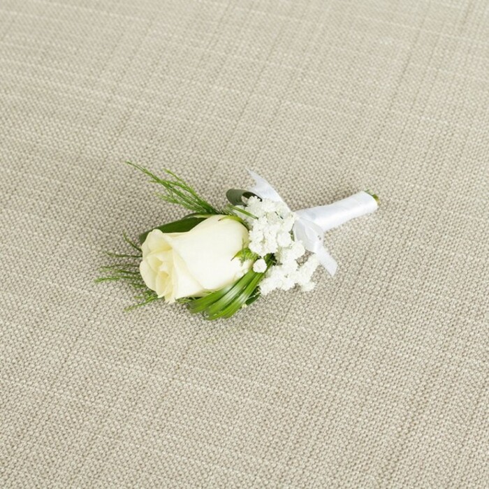 A refined single-rose boutonniere is displayed diagonally across a softly textured, light beige fabric that resembles natural linen, its fine weave and gentle crosshatch pattern evoking the relaxed elegance often seen at New Barnet weddings and receptions. The focal point is a single creamy white rosebud, its petals smooth, velvety, and still largely closed, with only a delicate curl at the outer edges hinting at the fullness to come. The centre of the rose remains hidden, lending a sense of purity and anticipation that suits a groom preparing to walk down the aisle at a local church near New Barnet Station or a nearby registry office. At the base of the bloom, hints of fresh green sepals provide a subtle contrast, reminding you that the flower has been cut and arranged with care not long before the event. Clusters of airy white baby's breath nestle to the right and slightly behind the rose, their tiny, star-shaped blossoms forming a gentle halo that lightens the composition and adds a soft, cloud-like texture. Interwoven with these delicate filler flowers are slender, rich green leaves and a few feathery, fern-like sprigs, thoughtfully placed to frame the rose without overwhelming it. The foliage introduces depth and dimension, making the small arrangement feel complete and well-balanced, as if it has been designed by an experienced local florist used to dressing lapels for celebrations across New Barnet and neighbouring Barnet Green spaces. The stems are carefully aligned and then tightly wrapped in a pristine white satin ribbon, the material smooth and slightly glossy, reflecting the soft, diffused light that falls across the scene. Just beneath the floral cluster, the ribbon is tied into a neat, symmetrical bow, the loops and tails perfectly proportioned to the size of the boutonniere, signalling meticulous craftsmanship and attention to fine detail. The lighting is gentle and even, casting minimal shadows and allowing each texture to be appreciated-the satin sheen of the ribbon, the delicate structure of the baby's breath, the subtle ridges on the rose petals, and the faint grain of the background fabric. Overall, the image conveys a sense of timeless, classical sophistication, ideal for illustrating formal wedding attire, groom's accessories, and the understated romance that many couples in New Barnet choose for their special day.