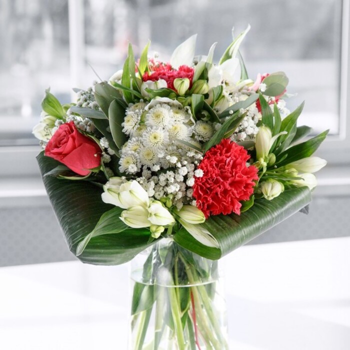 A luxurious red and white bouquet is captured here in soft, natural light, its colours glowing gently against a bright, understated backdrop that could easily be a New Barnet dining room or office space. The flowers are arranged in a tall, clear cylindrical glass vase, the water crystal clear and revealing a bundle of neatly cut green stems beneath the surface, speaking of early-morning conditioning and careful preparation. At the centre, several deep red roses unfurl in velvety spirals, their rich colour and flawless form giving a classic, romantic quality to the arrangement. Just in front, a large crimson carnation with deeply ruffled, densely layered petals provides dramatic texture, like a finely gathered piece of silk or lace. Around these bold red blooms are clusters of white and cream flowers: white alstroemeria with slender petals and subtle speckling at the throat, and a few elegant white lily buds still tightly wrapped, poised to open and gradually change the balance of the bouquet over time. Fine sprays of gypsophila (baby's breath) are threaded throughout, their tiny white blossoms creating a soft, cloud-like effect and adding a sense of celebration without overwhelming the structure. Embracing all of this colour is a generous frame of greenery. Broad, glossy dark green leaves are artfully twisted and wrapped around the base like a green collar, giving the bouquet a tailored, contemporary edge. Additional foliage in multiple shades of green-some with the silvery cast of eucalyptus, others pointed and upright-adds volume, texture, and a lightly herbal scent. The vase stands on a pure white surface, the background softly blurred so that nothing distracts from the flowers themselves. The mood is sophisticated yet welcoming, ideal as a centrepiece for an anniversary dinner near Oakleigh Park, a thoughtful Mother's Day surprise delivered across New Barnet, or a refined gesture of gratitude to brighten a client's reception area.
