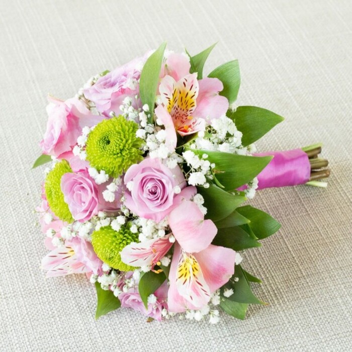 In this radiant bouquet, expertly arranged and photographed atop a pale, softly textured fabric reminiscent of New Barnet's artisan market stalls, every element reflects thoughtful local craftsmanship. At first glance, pastel pink roses-each with delicately layered petals-pattern the arrangement, emanating a subtle fragrance and timeless elegance. Interwoven with these, vibrant lime green button chrysanthemums catch the eye with their unique spherical forms and tactile texture, a lively contrast echoing the energy of the High Road. Pink alstroemeria, or Peruvian lilies, appear in gentle gatherings, their petals painted with hints of yellow and intricate dark markings for a dash of exotic refinement. Wispy white gypsophila adds a sense of buoyancy and dream-like softness, floating among the main blooms and lightening the arrangement's mood. Structural depth is provided by lush green foliage that frames the composition, as if borrowed from the tranquil green spaces surrounding New Barnet. The gathered stems are bound in a rich plum satin ribbon, tied with precision to balance vibrancy and elegance while leaving the raw cut ends artfully exposed. A gentle, diffuse light bathes the entire bouquet, enhancing the palette's romance and allowing each flower's unique texture to shine. Whether chosen as a bridesmaid's bouquet for a wedding celebration or simply as a heartfelt gesture of affection, this hand-tied arrangement connects the beauty of skilful floristry with the character and spirit of New Barnet community life.