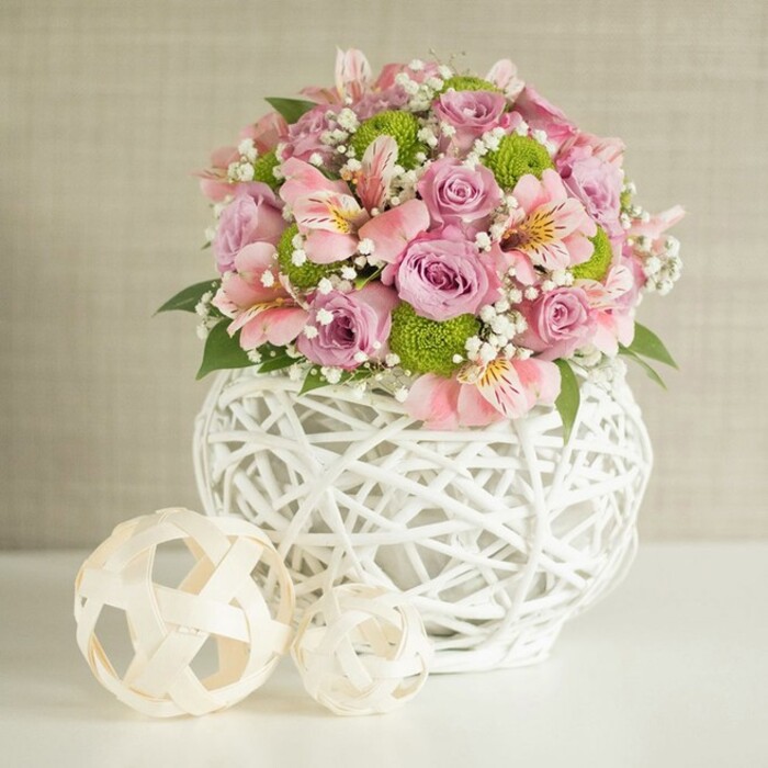 A lush and radiant wedding table centerpiece designed in New Barnet style, this voluminous floral arrangement blends contemporary artistry with classic romance. At its heart, layers of pastel pink and lavender roses unfold luxuriously, each petal softly illuminated by filtered light that recalls tranquil afternoons along Crescent Road. The roses are complemented by dainty stems of pink alstroemeria, their signature striping and subtle golden highlights adding intrigue. Sprightly lime green button chrysanthemums pop amidst the blooms, their texture and brilliance bringing to mind the manicured greenery of nearby parks. Echoes of white baby's breath weave throughout, providing ethereal lightness-a breath of fresh air akin to a window flung open on a gentle day in New Barnet. The arrangement is anchored in a distinctive white vase, its curving, open-weave lattice structure evocative of hand-crafted elegance. Two decorative spheres placed on the left side, crafted from pale woven reeds, reinforce the pastoral-modern aesthetic and draw the eye with their organic forms. The scene is framed by a calm, textured beige background, letting the pastel hues take center stage. Ideal for wedding receptions in local venues or as an elegant home accent, this composition exudes freshness, artistry, and a subtle nod to New Barnet's unique blend of tradition and innovation. Perfect for elevating celebrations or daily living across the community.