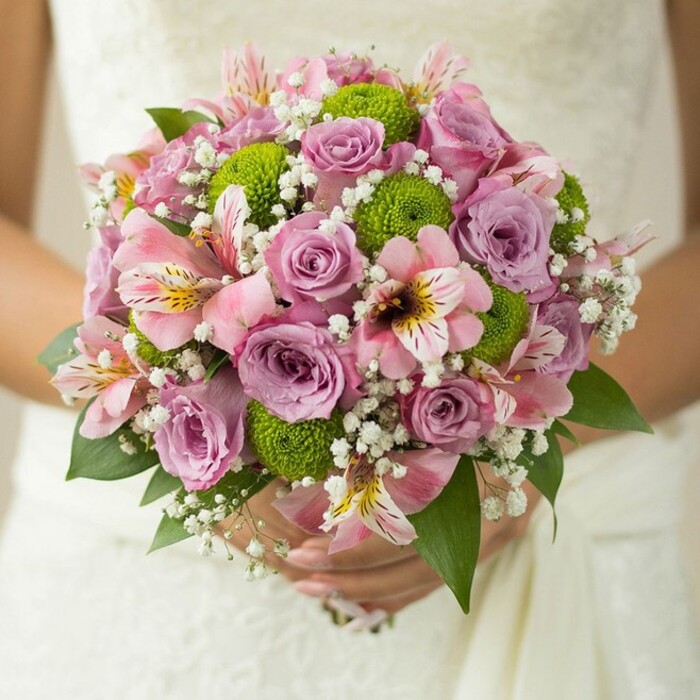 Glowing in soft natural light, this stunning bridal bouquet-expertly arranged by a local New Barnet florist-rests against the embroidered white fabric of a wedding gown, hands delicately embracing the stems at center frame. Its rounded, lush shape is alive with movement and contrast, blending tradition with fresh, cheerful touches. In its heart are romantic roses, some a gentle lavender-purple and others a sweet pink, petals dusted with traces of morning dew. Between these blooms, vibrant lime green button chrysanthemums introduce a playful pop, their spherical shapes reminiscent of the lively energy along New Barnet's bustling streets. Pink alstroemeria lilies, marked with dark streaks and sunny yellow throats, lend an exotic elegance and carry the scent of spring, recalling the floral stalls on Greenhill Parade. Wispy clouds of baby's breath weave through the arrangement, their tiny white florets accentuating the bouquet's volume and lending an ethereal softness. Rich, dark green leaves peek out at intervals, grounding the lighter tones and illuminating the flowers' vivid colors. The bride's hands cradle the bouquet with gentle reverence-a silver band glinting softly-signifying both commitment and anticipation. This composition exudes romance, sophistication, and modern flair, epitomizing the joy of a wedding day in New Barnet. Whether destined for a celebration on Station Road or an elegant event near Victoria Park, the bouquet encapsulates the beauty of local artistry and the everlasting promise of love.