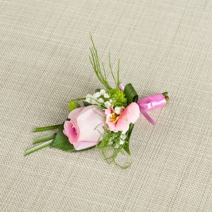 Resting at an angle across a subtly woven, light beige backdrop, this meticulously crafted boutonniere radiates the understated charm often found in New Barnet's community celebrations. A single, blush pink rose anchors the arrangement, its petals in a tight, graceful curl that hints at youth and hope. Nestled beside it, a paler blossom-possibly alstroemeria in peachy tones-unfurls with a hint of yellow and finely etched lines, lending intricate detail to the composition. Accents of snowy baby's breath drift amidst small, fresh green button-like mums, evoking textures of morning dew on local greenery in places such as Tudor Park. Diverse greenery, from slender grass-like fronds to flat, dark green leaves and even a neatly wrapped leaf at the base, provides structure and a lush backdrop-mirroring the balance of cultivated gardens and wild spaces unique to New Barnet. Everything is tied together by a striking fuchsia pink satin ribbon, whose glossy bow infuses a modern, festive note. Gentle, natural light bathes the scene, making each hue and sheen stand out against the tactile fabric, whose crosshatch threads amplify the freshness of the blooms. This boutonniere, ideal for weddings or landmark occasions, speaks to both tradition and creativity-a refined accessory crafted by local hands, imbued with the welcoming spirit of New Barnet.
