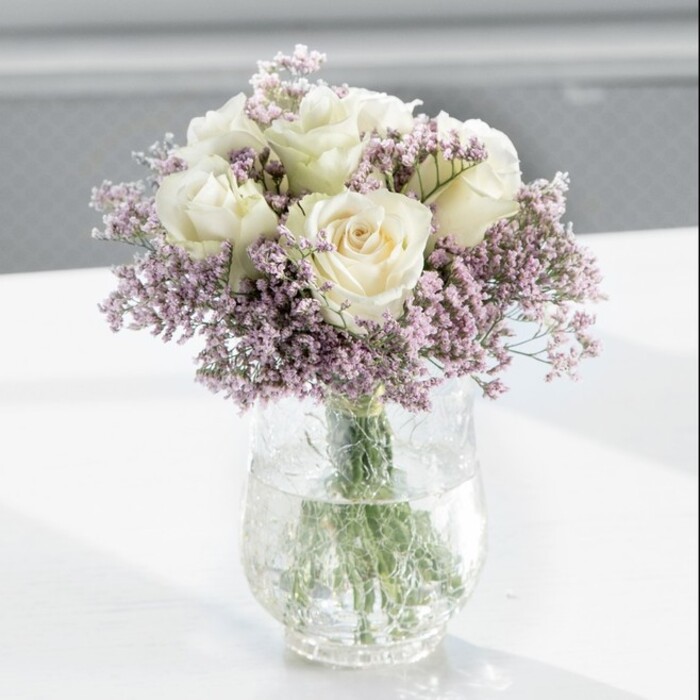A serene and elegant bouquet of creamy white roses framed by lilac-toned statice is the clear focal point of this image, positioned proudly in a tall, cylindrical glass vase on a pristine white surface. The roses are abundant and evenly spaced, forming a classic, rounded shape that feels both considered and natural, like something you might see gracing a sunlit bay window in a New Barnet home overlooking Victoria Park. Each rose reveals layer upon layer of soft, velvety petals, opening from a gentle, warm centre to slightly ruffled edges, creating a sense of depth and quiet luxury. Interwoven among these statement blooms are plentiful sprays of fine statice flowers in light lavender-purple shades; their tiny clustered petals and slender stems introduce a feather-light texture, a delicate haze that wraps around the roses and softens every outline. Through the glass, the straight green stems are clearly visible, gathered tidily and submerged in fresh, clear water, which adds a cool, reflective element to the composition. Natural daylight streams in from the side, bathing the bouquet in a tranquil glow, touching the rose petals with soft highlights and causing subtle reflections on the glossy tabletop. The background is intentionally blurred, suggesting a calm, contemporary interior with pale walls and perhaps a large window, without drawing attention away from the flowers. The overall mood is peaceful and refined, perfect for marking a gentle celebration, sending condolences to a family home off Barnet Hill, or welcoming a new baby with understated grace. The design feels like the work of a careful local florist who understands how to express emotion through quiet, timeless beauty.