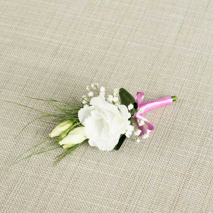 A finely composed boutonniere, photographed from a slightly elevated angle, rests on a warm, oatmeal-toned fabric background whose subtle woven texture suggests natural linen laid out on a table in a New Barnet townhouse. At its centre, a single white lisianthus flower is fully open, its petals soft and gently frilled, layered in concentric waves that form a rounded, romantic blossom. The bloom's creamy white tone glows softly against the neutral fabric, giving it an almost luminous presence. Just to the side and slightly below this focal flower, two elongated lisianthus buds lean together, their pale green and ivory tips still closed, capturing the poetic sense of a story just beginning-much like a couple preparing for their vows at a New Barnet church. Around the main flower and buds, light, cloudlike sprays of white gypsophila cluster in small, airy tufts, adding a delicate, lace-like texture that softens every edge. Supporting the flowers is a mix of greenery: a smooth, darker green leaf sits discreetly behind the lisianthus, while several fine, grass-like blades extend gracefully toward the left side, introducing movement and a natural, just-picked-from-the-garden feeling. The stems are carefully aligned and tightly wrapped at the bottom with a vibrant band of fuchsia pink satin ribbon, tied into a small but confident bow whose bright, modern colour provides a joyful counterpoint to the classic white blooms and foliage. The even, gentle lighting reveals each detail-the silky petals, the tiny gypsophila clusters, the satin sheen of the ribbon-forming a refined lapel piece perfect for grooms, ushers, or prom-goers in New Barnet who appreciate understated elegance with a playful pop of colour.
