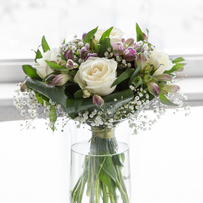 A graceful, thoughtfully composed sympathy bouquet is captured here, standing in a clear cylindrical glass vase that showcases the straight, green stems gathered neatly together. The flowers themselves form a rounded, lush dome of whites, purples, and greens, gently lit as if by natural light streaming through a New Barnet window. Plush white roses are the main focus, their petals layered like soft satin, edges just beginning to curl outward in full, open blooms. These roses are nestled among slender stems of alstroemeria, whose trumpet-shaped flowers display subtle gradients of colour from pale lavender and soft violet to a fresh, almost lime-tinged green at the throat, reminiscent of early blooms in nearby Tudor Park. Some alstroemeria blossoms have opened wide, revealing delicate markings inside each petal, while others remain closed buds, adding height and quiet anticipation to the design. Fine sprays of baby's breath, with countless tiny white flowers, drift through the arrangement like a light mist, softening the stronger shapes and bringing a touch of airiness to the clustered blooms. Around and beneath the flowers, rich green foliage in varying shapes and textures creates depth and structure, cradling the roses and alstroemeria while keeping the bouquet visually balanced. The background is bright and softly blurred, a field of pure white that allows the subtle colours of the bouquet to stand out clearly, without distraction. The whole arrangement conveys a mood of calm support and sincere condolence, an elegant floral gesture that would feel at home in a quiet New Barnet living room, at a memorial service, or beside a framed photograph of someone dearly missed.