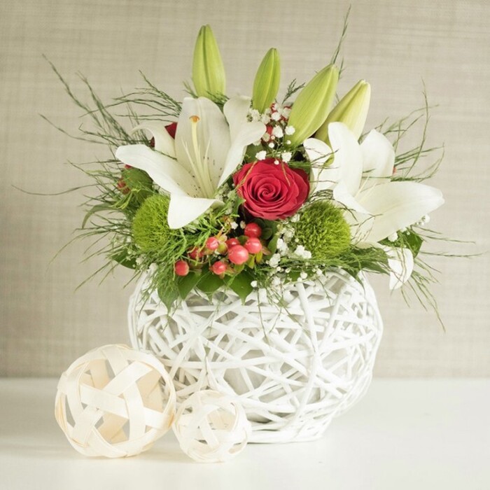 On a clean white tabletop, a generously styled floral arrangement stands as a refined focal point, reminiscent of the elegant table decorations chosen for New Barnet weddings and family celebrations. The flowers are gathered into a round, sculptural vase made of white woven strands that cross and loop to form an open, basket-like lattice, giving the container a light, airy presence rather than heavy solidity. From this textured base rises a lush bouquet dominated by white lilies, their petals opening outward in broad, star-like shapes. Some blooms are fully open, revealing soft, creamy interiors and well-defined yellow stamens dusted with pollen, while others remain mostly closed, slender green buds reaching upwards and suggesting ongoing freshness, as if the display were prepared that very morning by a local florist. Among the lilies, deep red roses provide dramatic contrast: one especially striking rose sits near the centre front, its tightly coiled petals forming a near-perfect spiral, drawing the eye like a jewel set against ivory fabric. Spherical tufts of bright green pompon flowers, most likely dianthus or small chrysanthemums, tuck into the spaces between the larger stems, adding a cushiony texture and vibrant pop of colour that keeps the design feeling youthful and vivid. Fine sprays of white baby's breath drift throughout, softening the contrasts and offering a light, cloud-like layer between the stronger shapes. Red hypericum-style berries and an array of greenery-from feather-soft, trailing fronds to broader, deeper green leaves-create dimension and a sense of natural abundance, evoking walks through the leafy streets around Oakleigh Park. To the left of the main vase, two decorative woven spheres in pale, natural wood tones echo the open weave of the container and give the scene a cohesive, curated feel. Behind the arrangement, a softly textured beige-grey wall keeps the background quiet and neutral, allowing every petal, leaf, and woven strand to stand out, resulting in a centrepiece perfectly suited to a wedding table or intimate celebration in New Barnet.