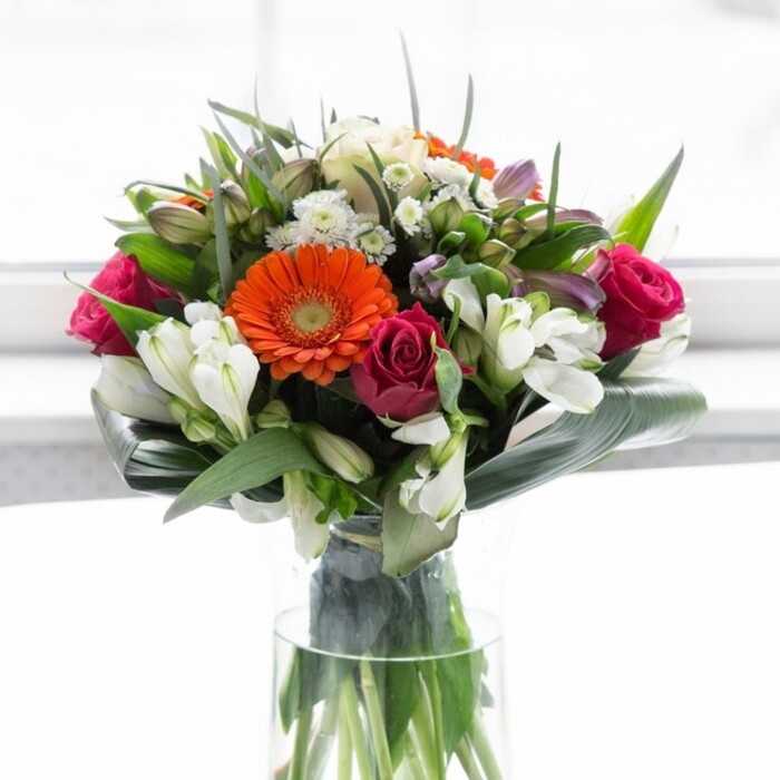 This exquisite floral arrangement is a testament to New Barnet's love of craft and celebration, elegantly presented in a clear glass vase that showcases every fresh green stem. The heart of the bouquet features peach and orange gerbera daisies, their wide petals and sun-burst centers recalling the early morning glow on New Barnet streets. Nestled between these cheerful flowers, velvety fuchsia roses exude depth and timeless sophistication, their spiral forms drawing the eye into the bouquet's center. Flanking the main blooms, soft white alstroemeria and dainty spray roses lend an ethereal, feathery touch, reminiscent of the gentle haze over Oak Hill Park on a spring morning. Flourishes of pale pink and tiny white fillers contribute layers of texture and a sense of lush abundance. Underpinning the blooms are boldly curved, glossy aspidistra leaves, wrapping protectively around the base and accentuating the arrangement's rich, rounded silhouette. Every space between stems is filled with fresh greenery-each leaf and sprig chosen to bring contrast and natural harmony. Bathed in calm, diffused daylight, this bouquet feels alive with promise and warmth. It is perfectly suited for marking joyous milestones, expressing gratitude, or welcoming a new chapter, channeling the inviting spirit of New Barnet's high streets, markets, and neighbourhood celebrations.