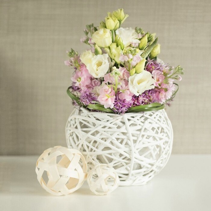 A luminous floral arrangement, crafted by the skilled hands of New Barnet florists, commands attention with its elegant interplay of pastel hues and intricate textures. At the arrangement's heart, layers of snowy white and soft yellow lisianthus swirl together, their ruffled petals unfolding with quiet grace beside slender stalks of pink matthiola, which reach gently upward, imparting movement and lightness. Underpinning this floral canopy, clusters of rich purple aster or similar filler flowers fan outward, their vibrant color grounding the bouquet and lending dimension. Lush green leaves surround the blooms, creating a crisp rim that frames the lively abundance. The entire bouquet sits snugly in a contemporary globe-shaped vase, its white painted rattan latticework airy and sophisticated-a testament to modern artistry found along East Barnet's creative thoroughfares. To one side, a pair of decorative orbs-each handwoven from pale natural fibres-rest serenely, reflecting both the vase's material palette and the borough's appreciation for craft traditions. The composition is illuminated by soft, ambient daylight that dances lightly across a neutral greige backdrop, suggesting a well-loved home or stylish New Barnet venue. Overflowing with freshness and intentional design, this table centrepiece conjures a mood of peaceful celebration, ideal for intimate spring weddings, milestone anniversaries, or simply adorning a local dining table with a breath of seasonal joy.
