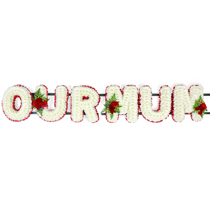 This photograph shows a dignified floral letter tribute spelling out OUR MUM, arranged in a straight line across a bright, uncluttered white background that gives the design a calm, contemplative presence. Each capital letter is formed on a solid frame and completely blanketed with small white chrysanthemum florets, placed petal to petal so that the surface appears luxuriously full and cushion-like. Their fresh, clean white colour conveys purity and peace, while the clustered heads create a pleasing, slightly domed texture that suggests softness to the touch. Encircling each letter is a continuous border of deep red blooms, probably spray carnations, carefully wired and tucked into place to create an even, ribbon-like edging. This bold red frame offers a striking contrast and ensures the shapes of the O, U, R, and the two Ms are clearly defined even from a distance, as they might be when viewed at a funeral service in one of New Barnet's local chapels. The letters are discreetly connected by a dark green metal or plastic bar along the back, barely visible beneath the flowers but providing essential support. Adding an extra layer of sentiment, three small groupings of deep red roses, paired with rich green foliage and delicate fern fronds, are nestled between the letters: one between O and U, one between U and the first M, and one accenting the end of the final M. These rose sprays bring depth, movement, and emotional warmth, their velvety petals hinting at gentle fragrance and long-held memories. Created in the thoughtful style local families around New Barnet often request, this OUR MUM tribute feels deeply personal, a quiet yet powerful expression of love and gratitude for a mother who is dearly missed.