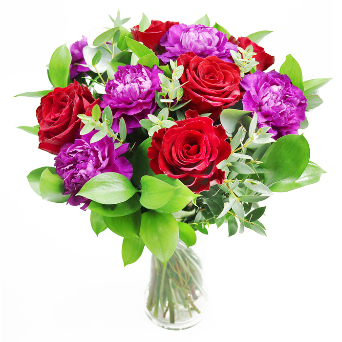 This arrangement presents a refined conversation between colour and texture: deep red roses and vibrant purple peonies gathered with lush greenery in a simple clear vase, as if styled for an elegant New Barnet townhouse just off Station Road. The roses are richly saturated, their velvety petals spiralling outward in neat, generous whorls that speak of timeless romance and heartfelt emotion. Purple peonies nestle among them, their blossoms rounded and full, each one a cushion of intricate, softly ruffled petals that seem to billow like silk. Around these focal flowers, a generous skirt of foliage provides depth and structure. Glossy green leaves arc outward, framing the blooms and adding a sense of movement, while slender stems of eucalyptus thread through the design, their silvery-green discs lending a contemporary, airy touch. The clear glass vase is understated but purposeful, revealing the straight, cleanly cut stems bundled together in tidy lines, recalling the well-kept gardens and tree-lined avenues near New Barnet's Hadley Green. Gentle, natural light falls across the bouquet, picking out subtle variations in the reds and purples and casting soft shadows on the surface beneath the vase. You can almost imagine leaning in to breathe the perfume: the rich, familiar scent of roses, softened by the faintly cool, aromatic note of eucalyptus and the freshness of just-cut foliage. This is the kind of bouquet that feels equally at home as an anniversary centrepiece, a dramatic birthday surprise, or a meaningful gesture of admiration delivered to a doorstep anywhere across New Barnet.