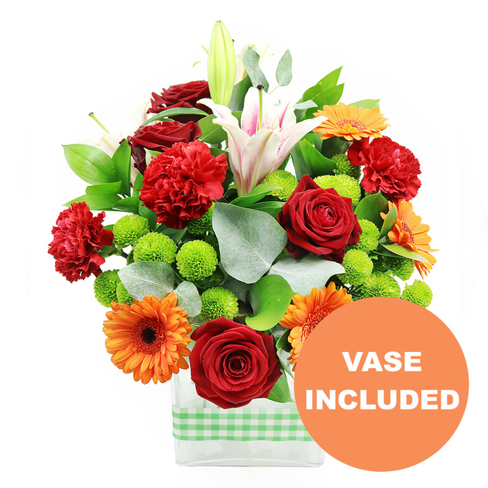 A richly packed, mixed-colour bouquet sits proudly in the centre of a white background, its vibrant reds, oranges, whites, and greens forming a lively focal point that feels perfectly at home in New Barnet's blend of suburban calm and city energy. The flowers are gathered into a clear, rectangular glass vase, its clean lines lending a modern edge while allowing the green stems to be visible, stacked and criss-crossed in an orderly grid. Encircling the lower portion of the vase is a light green and white gingham ribbon, a simple decorative band that gives a homely, rustic note, like something you might see in a stylish café near New Barnet station. At the heart of the arrangement, several deep red roses are in full, luxurious bloom, each petal curling softly over the next to create that classic, romantic rose shape. They are joined by frilled red and orange carnations, which bring a ruffled, almost cloud-like texture that softens the strong colours around them. Bright orange gerbera daisies punctuate the display with their wide, flat petals radiating outward from dark centres, creating bold discs of colour. Rising higher, white Oriental lilies offer elegance and movement; some are fully open, revealing pink-tinged throats and dusted stamens, while others remain in smooth green buds, suggesting fresh blossoms still to come. Between the larger focal flowers, tight clusters of lime green pompom chrysanthemums bubble up, adding a refreshing, almost zesty visual note. The whole bouquet is framed by a generous mix of glossy dark green foliage and matte silver-green eucalyptus sprigs, giving structure, depth, and a slightly aromatic feel. In the lower right corner, a bright orange circular graphic partially overlaps the vase, clearly declaring VASE INCLUDED in crisp white capital letters, making it clear this is a complete, ready-to-display floral gift ideal for same-day delivery to homes and offices across New Barnet.