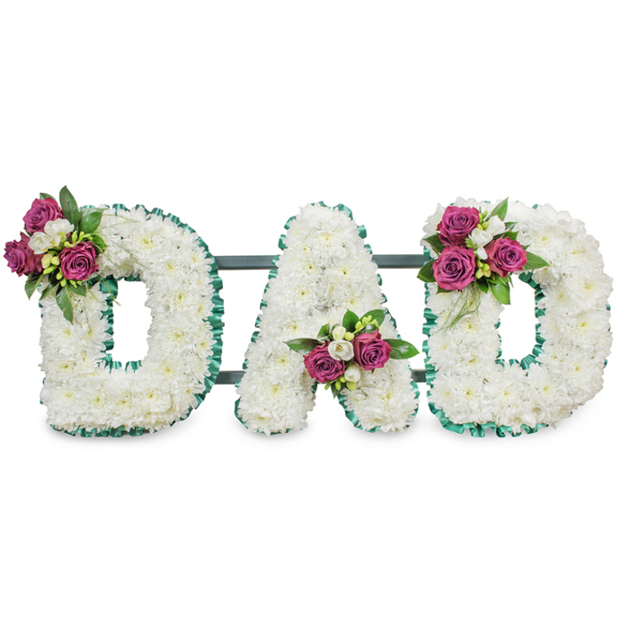 This memorial lettering spells DAD in substantial, capital forms, each letter composed of hundreds of pure white chrysanthemum florets that create a soft, pillowy texture and generous depth against a clean white backdrop. The craftsmanship is evident in the way each bloom is nestled close to its neighbour, producing uninterrupted contours that read clearly from a distance. A ruffled emerald-green ribbon crisply traces the outer and inner edges of every letter, its vivid hue framing the white field and lending a traditional, respectful accent. Each letter is embellished with a compact posy of deep magenta roses - some tightly coiled, some beginning to unfurl - mingled with small white freesia-like sprays and rich green foliage, placed to form a pleasing visual rhythm: an upper-left cluster on the first D, a centered V cluster on the A, and a mirrored upper-right cluster on the final D. Behind the letters a subtle dark green bar suggests a freestanding frame for display at a funeral or cemetery service. The scent is a soft combination of rose sweetness and fresh green stems under the clean chrysanthemum note, and the overall atmosphere is one of loving respect and quiet farewell. As Flowers New Barnet, serving homes and churches around New Barnet station, Brunswick Park and the A1000 corridor, we craft these bespoke tributes to reflect personal stories and steady remembrance for a dearly missed father.